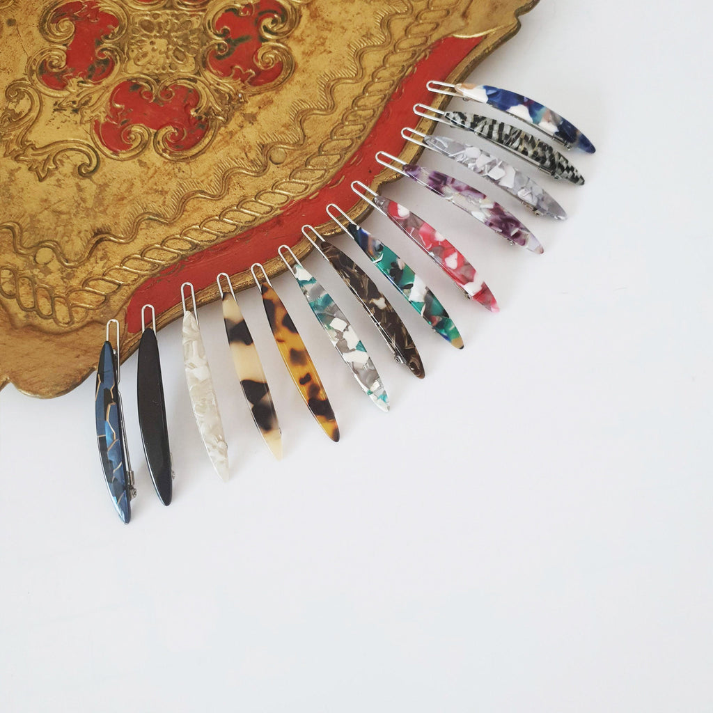 Cellulose acetate mini slim oval bobby pins in tortoise shell and marble patterns. 2.3 inch metal snap clips for women's side bangs. Minimalist Korean hair accessory. A collection of twelve colorful, patterned, plastic hair clips. The clips are arranged in a row, with the tips of the clips pointing to the right. The clips are all different colors and patterns, including black, white, brown, red, blue, and green. The clips are all made of a translucent plastic material.