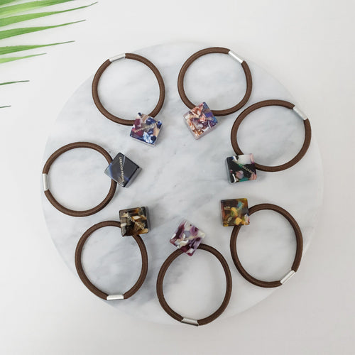 Cellulose acetate cube hair tie in tortoise shell and marble patterns. Geometric square ponytail holder. Modern Korean hair accessory. Six brown hair ties with square shaped charms. The charms are made of clear plastic with a colorful marbled pattern and gold lettering that reads 'CREATION GRACE'. The charms are attached to the hair ties with a silver metal ring.