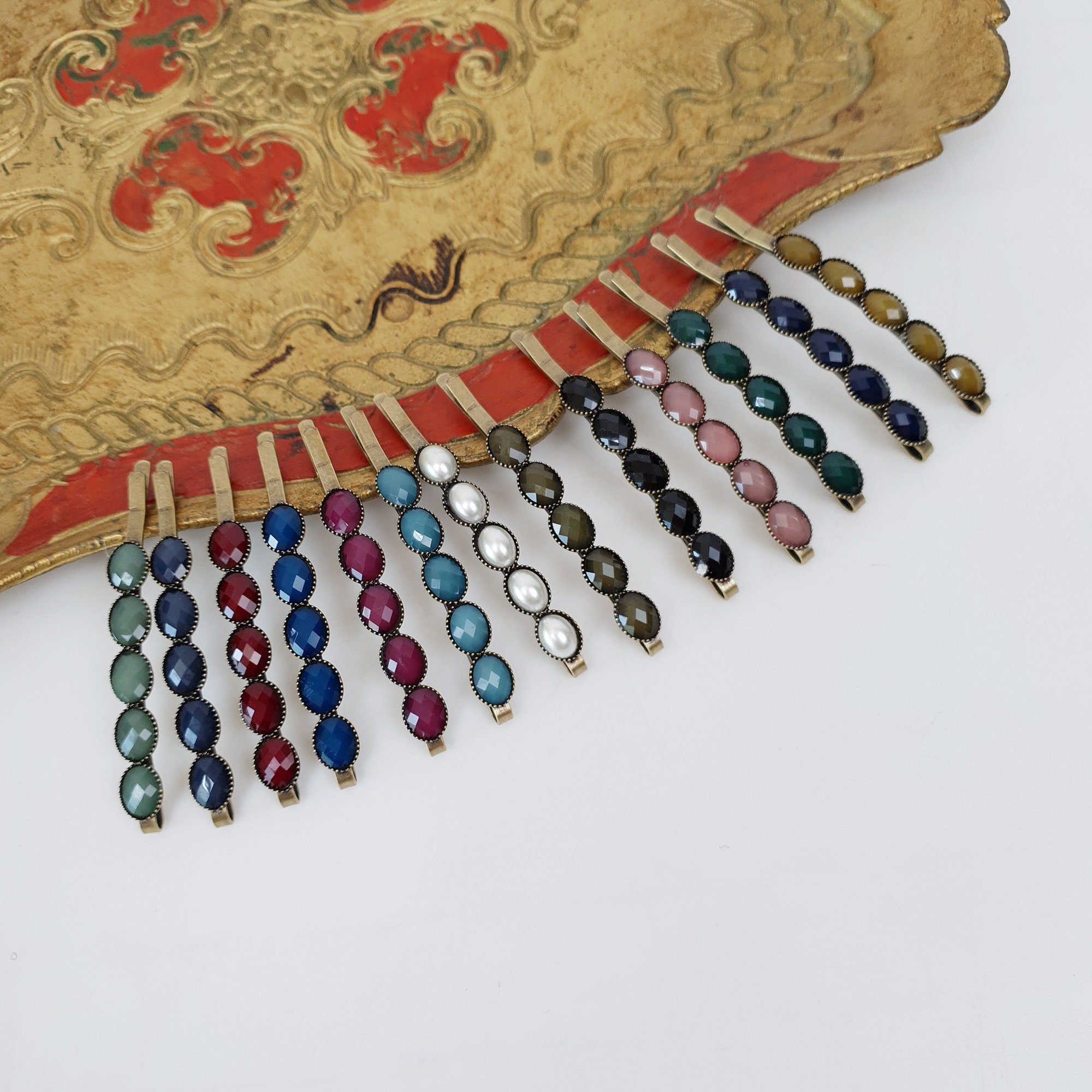 Rectangular vintage style formica hair slide with gold tone brass base. Bold straight line bobby pin in antique colors for styling side bangs or loose strands. A set of 14 hair clips with different colored rhinestones. The clips are gold colored and have five rhinestones each. The colors of the rhinestones are teal, blue, burgundy, pink, green, and white.
