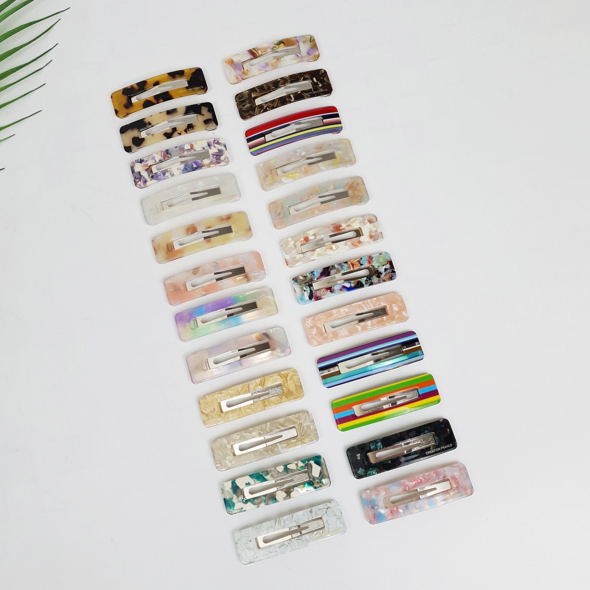 Rectangular cellulose acetate snap clip with cut-out center. Multicolored marble and tortoise pattern hair slide for side bangs, 90s retro style accessory. A collection of twenty hair clips in various colors and patterns. The clips are made of plastic and have a rectangular shape. The patterns include stripes, floral, and abstract designs. The clips are arranged in two rows of ten, with the top row slightly offset from the bottom row.