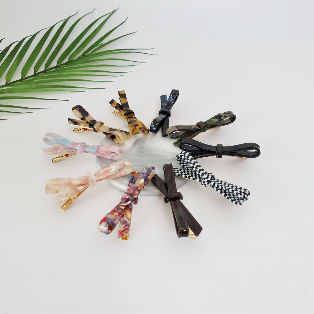 Cellulose acetate ribbon bow hair clip in tortoise shell and marble patterns. 2.8 inch alligator barrette for women's side bangs. Cute Korean fashion accessory. A collection of ten colorful hair clips with bow designs. The clips are arranged in a circle on a white surface. The bows are made of different materials, including plastic, fabric, and metal. The colors of the bows include black, white, brown, green, pink, and blue.
