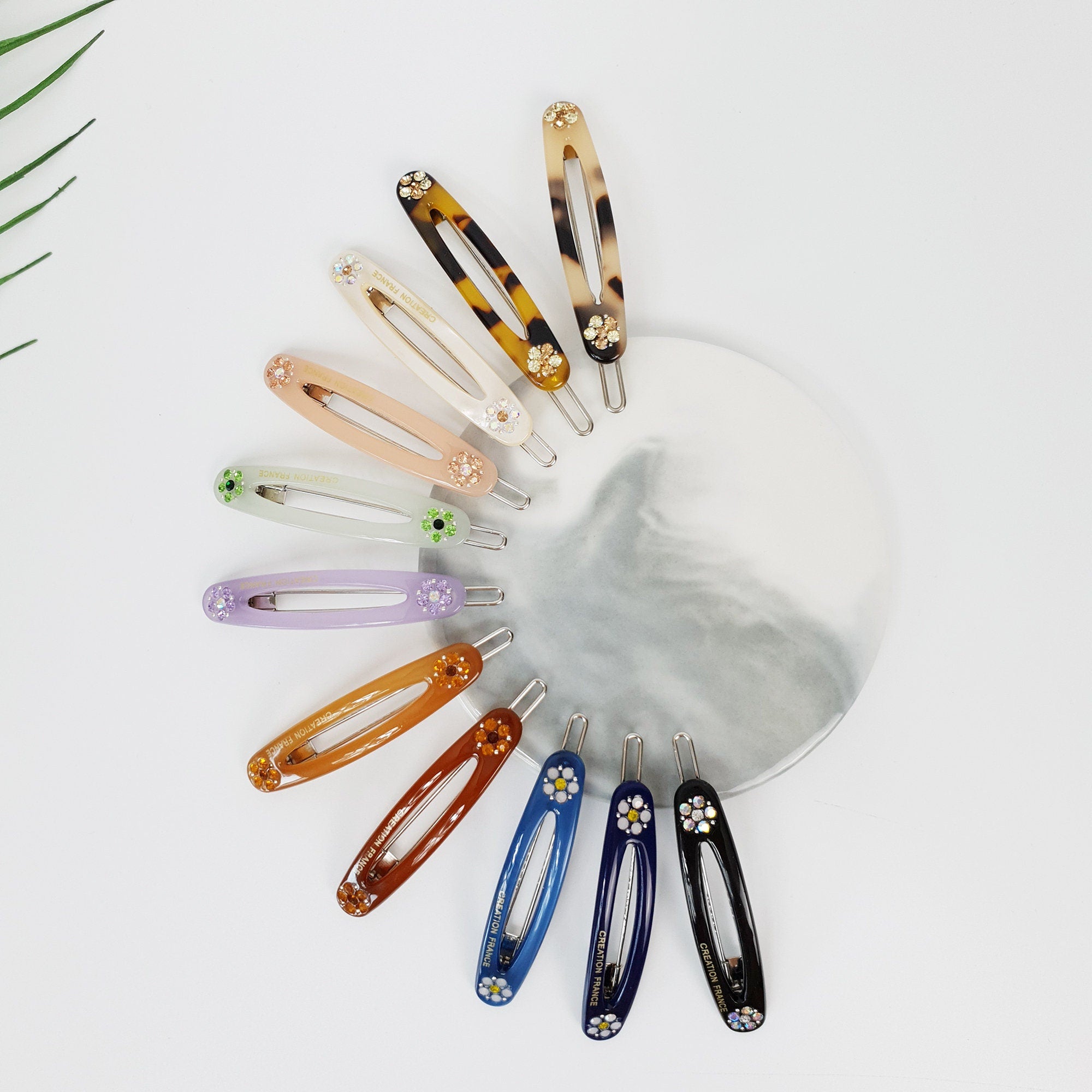 Cellulose acetate oval snap hair clip with rhinestone flower accents. Small tortoise shell and pastel barrette for bangs or side hair styling. A collection of twelve colorful hair clips with flower embellishments. The clips are arranged in a fan shape on a white surface. The clips are made of plastic and have a variety of colors, including purple, blue, brown, and beige. The clips have a small flower design on the top and a metal clasp on the bottom.