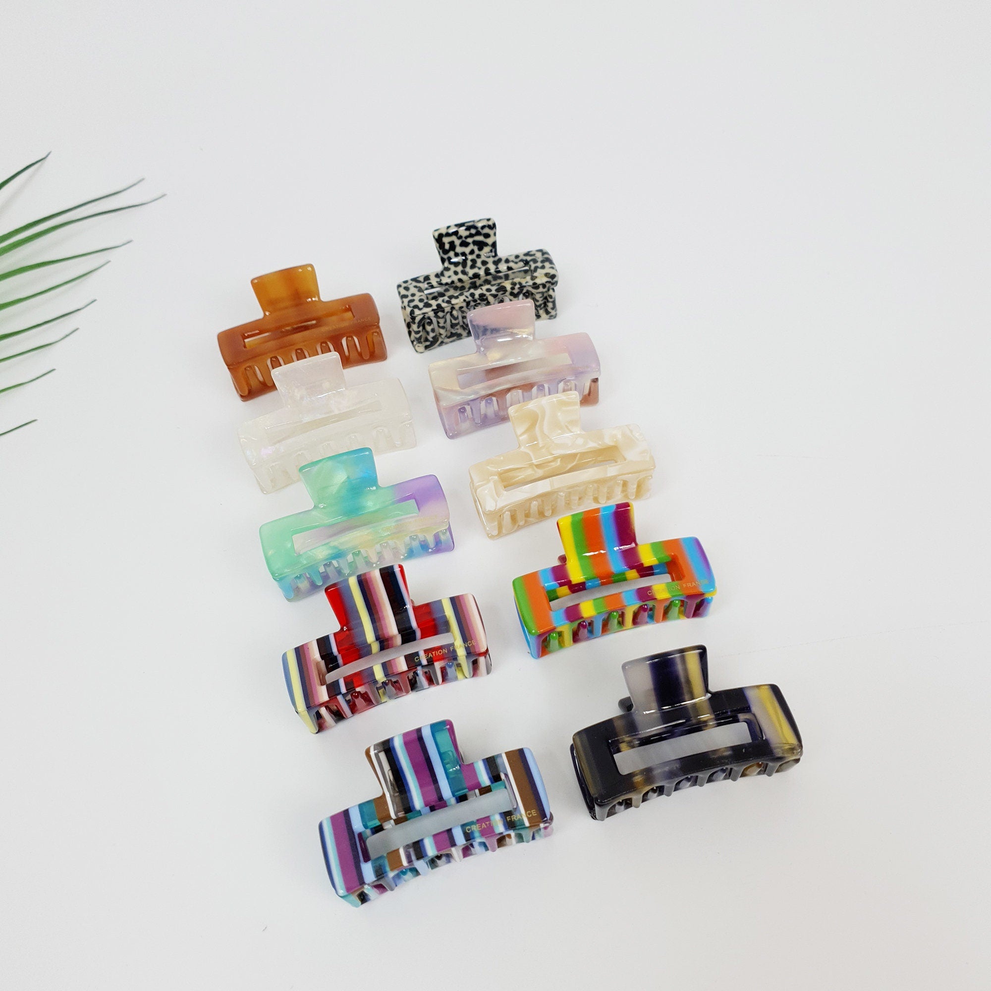 Cellulose acetate mini rectangular hair claw clip. 1.8 inch small jaw clip for bangs in tortoise shell, stripes, and aurora patterns. Cute geometric hair accessory for women.  A set of ten hair clips in various colors and patterns, including brown, black, white, pink, green, blue, red, yellow, and orange. The clips are made of plastic and have a claw-like design. The clips are arranged in a row on a white surface.