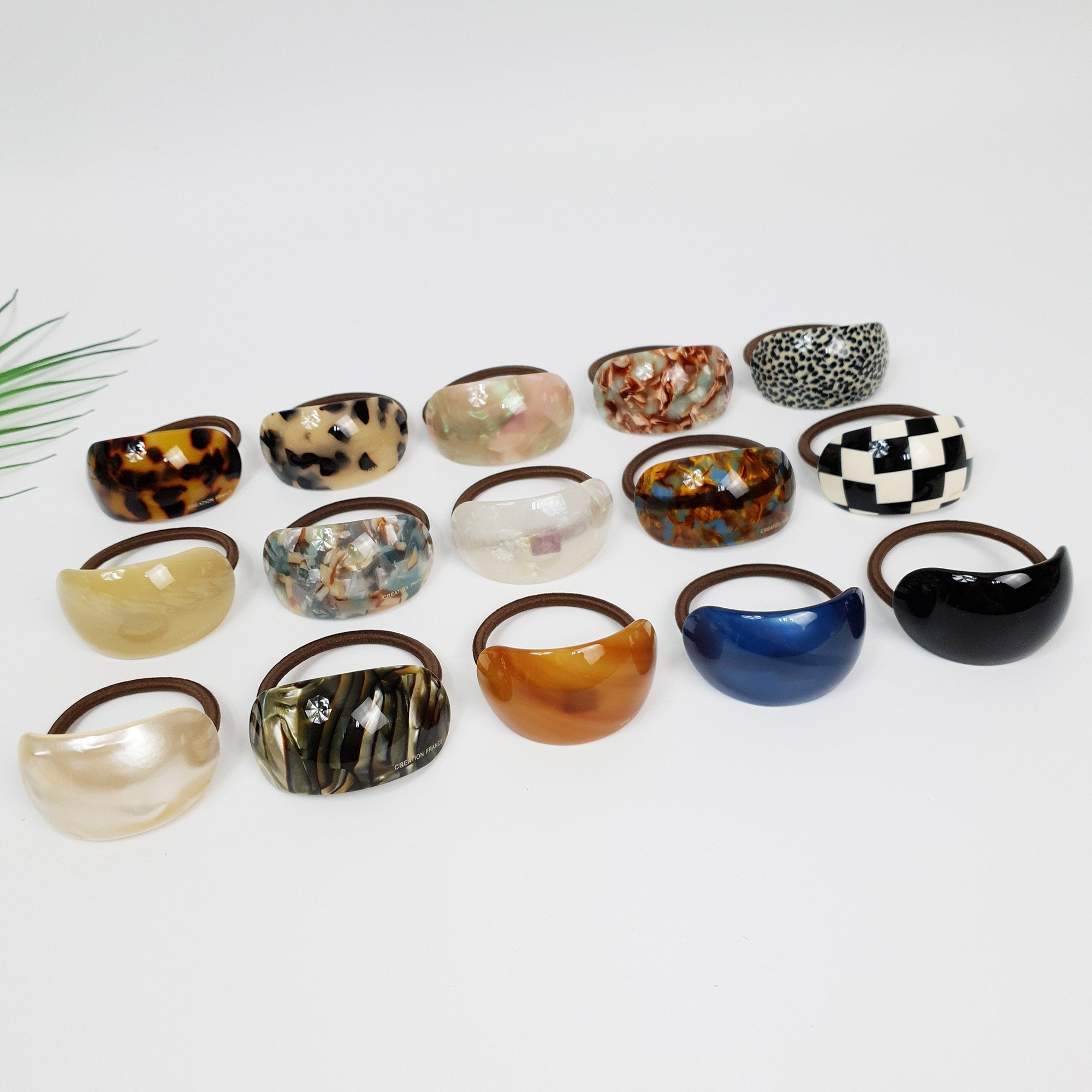 Cellulose acetate hair cuff with curved ponytail holder design, featuring tortoise shell, marble, and solid patterns for women. A collection of 15 hair scrunchies in various colors and patterns. The scrunchies are made of a hard plastic material and have a smooth, glossy finish. The scrunchies are arranged in a semi-circle on a white surface.