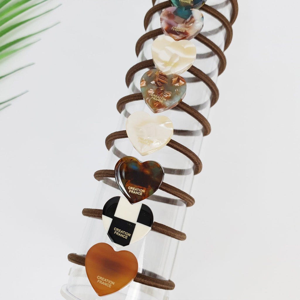 Small cellulose acetate heart charm elastic in brown and beige patterns. Dainty ponytail holder for women and girls. Glossy Korean accessory. A set of eight brown heart-shaped hair ties with a gold 'CREATION FRANCE' logo on each. The hair ties are arranged in a vertical stack on a clear glass tube.