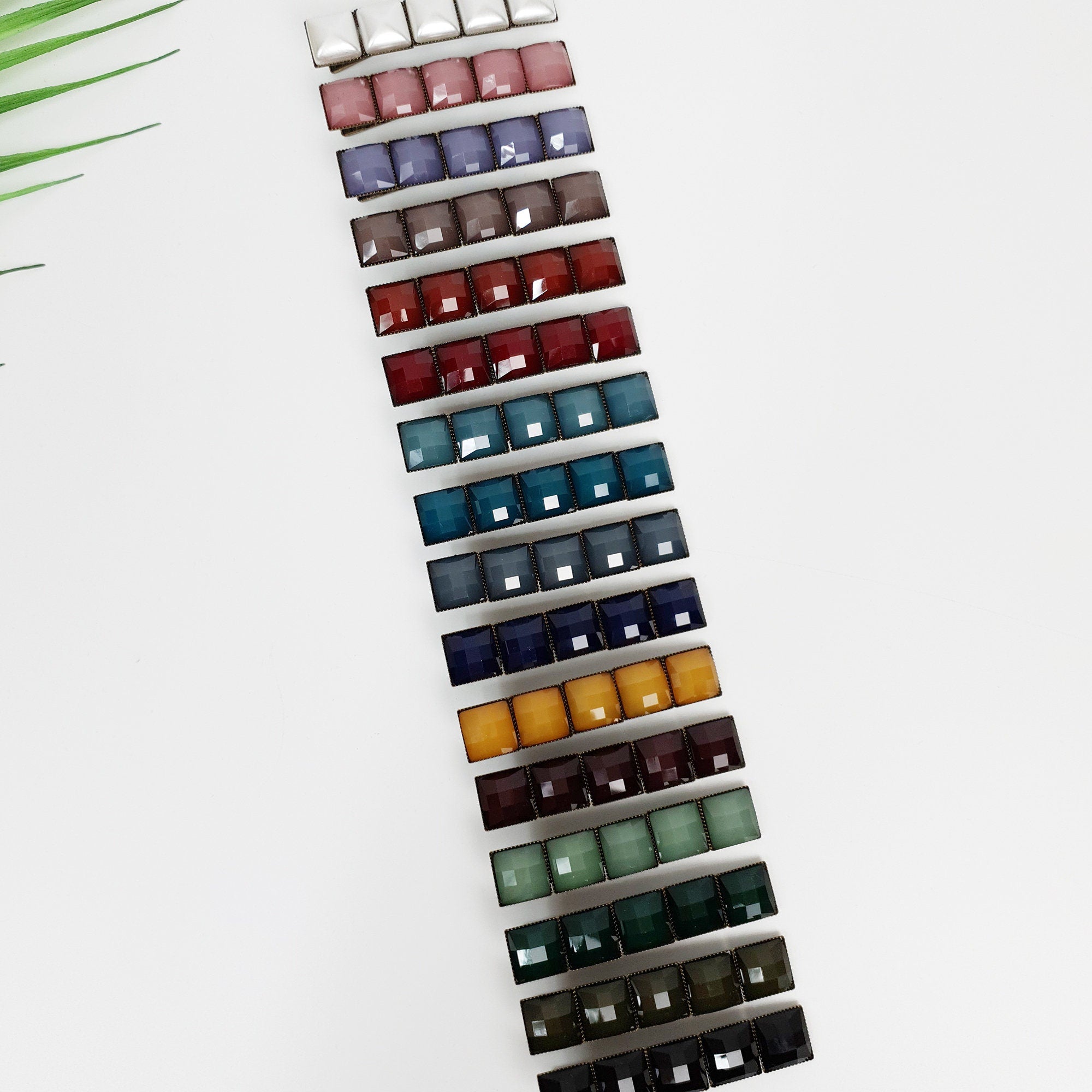 Vintage style square tile hair clip with antique brass finish. 2.4 inch mosaic alligator clip for side bangs and retro hairstyles. A set of 16 hair clips with square-shaped gems in various colors, including white, gray, brown, red, blue, green, and yellow. The clips are arranged vertically in a row, with the white gem at the top and the yellow gem at the bottom.