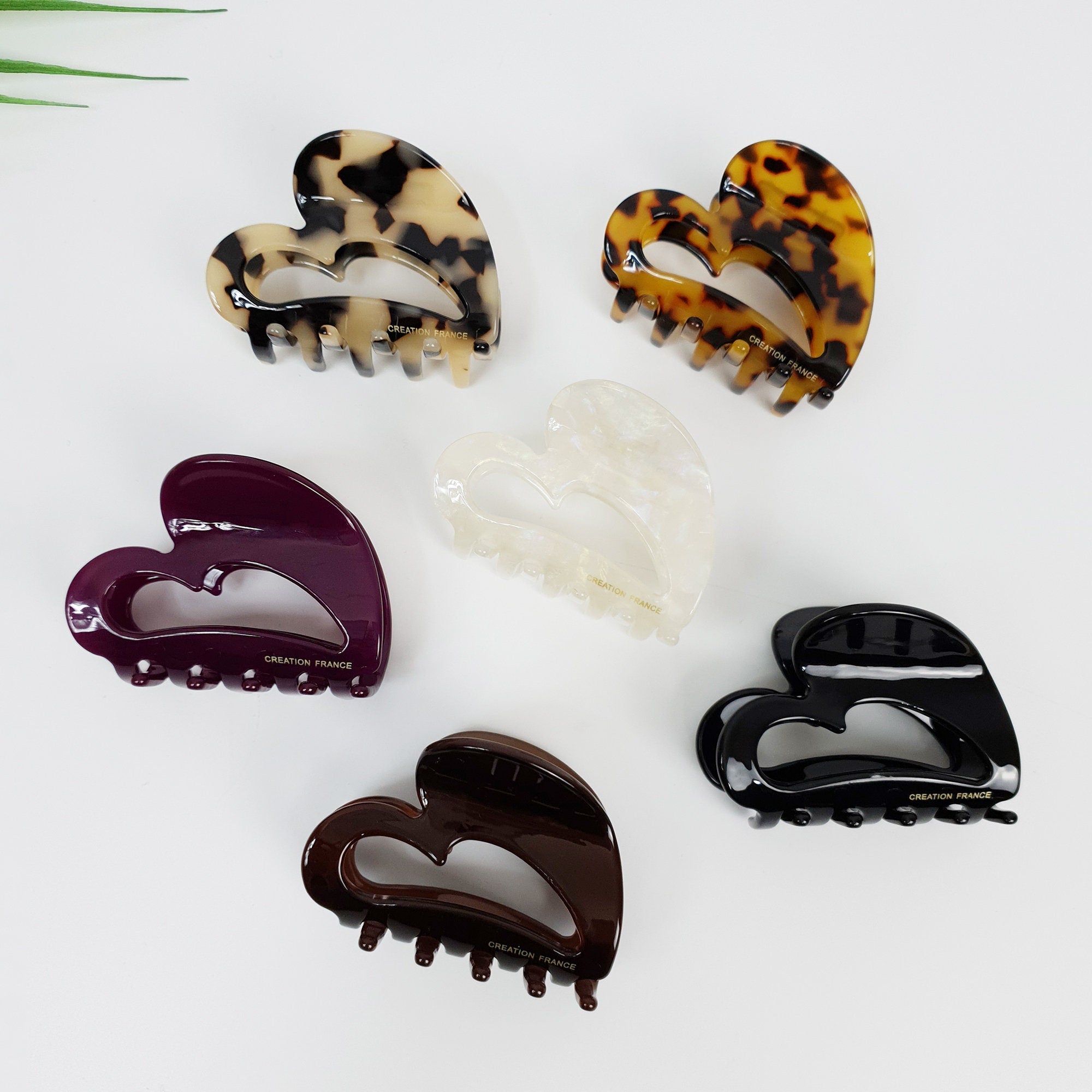 Cellulose acetate heart shaped hair claw clips in tortoise shell and solid colors. Medium 2.6 inch jaw clips for half up hairstyles. Five heart-shaped hair clips in various colors including black, brown, white, and tortoise shell. Each clip has a gold inscription that reads 'CREATION FRANCE'.