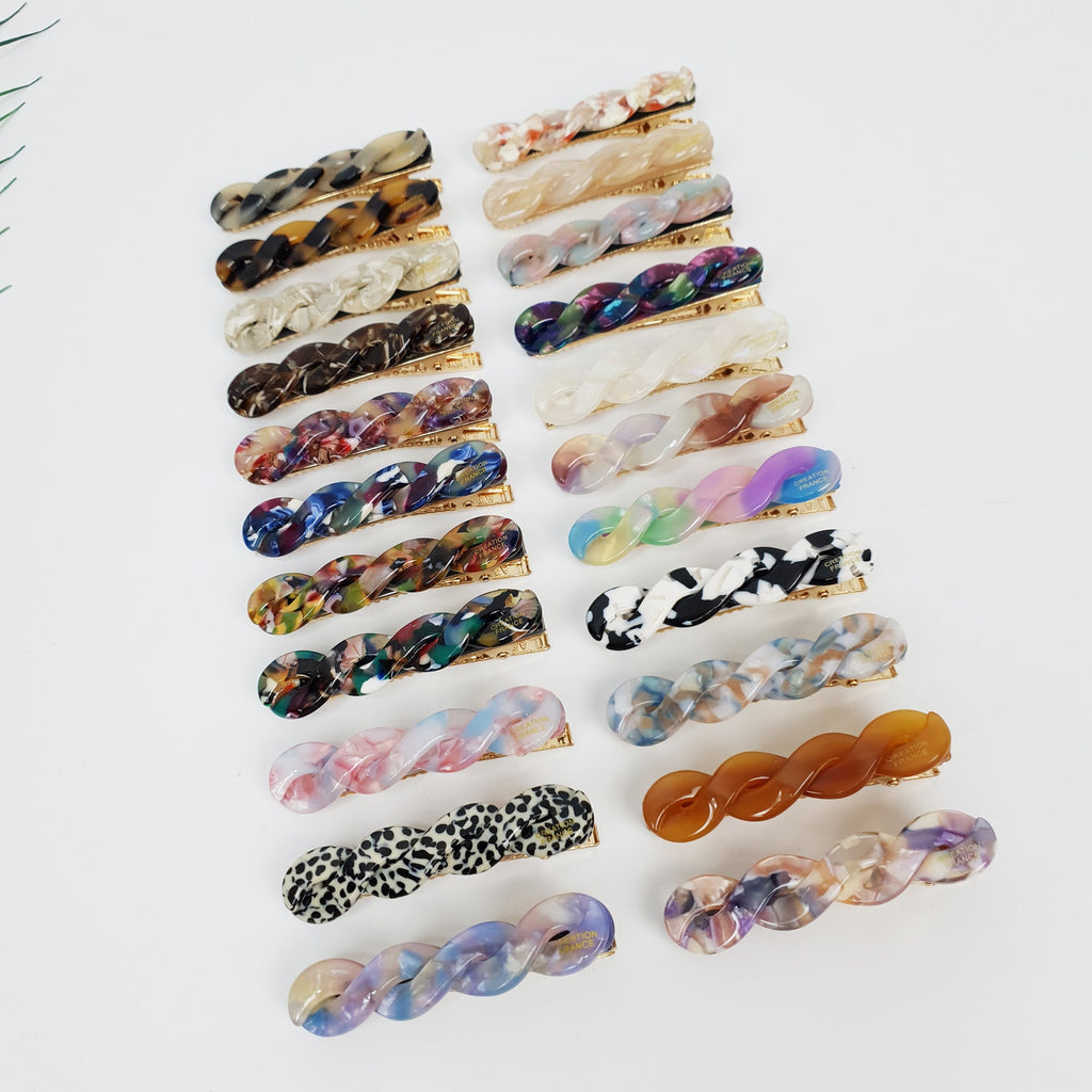 Cellulose acetate marble twist mini hair clip on model. 2.6 inch thin braided alligator barrette for women's side bangs. Minimalist Korean hair accessory. A collection of 18 assorted hair clips with a twisted design. The clips are made of plastic and have a variety of colors and patterns, including brown, white, black, pink, blue, green, and multi-colored. The clips are arranged in rows on a white background.