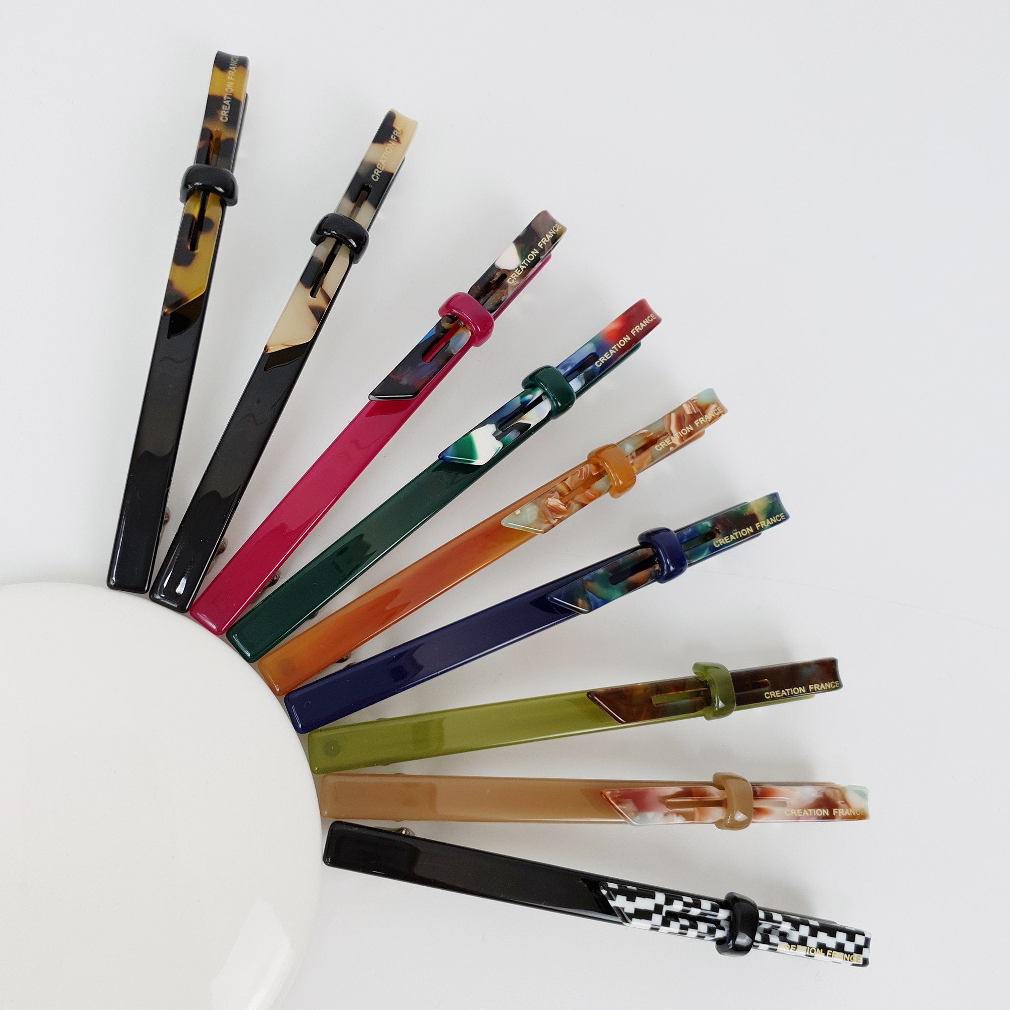 Cellulose acetate long skinny hair barrette in tortoise shell and solid colors. Slim 4.3 inch automatic hair clip for ponytails.  A set of nine colorful hair clips with a black and white checkered pattern on the end. The clips are made of plastic and have a variety of colors, including red, green, blue, orange, and brown. The clips are all labeled 'CREATION FRANCE'.