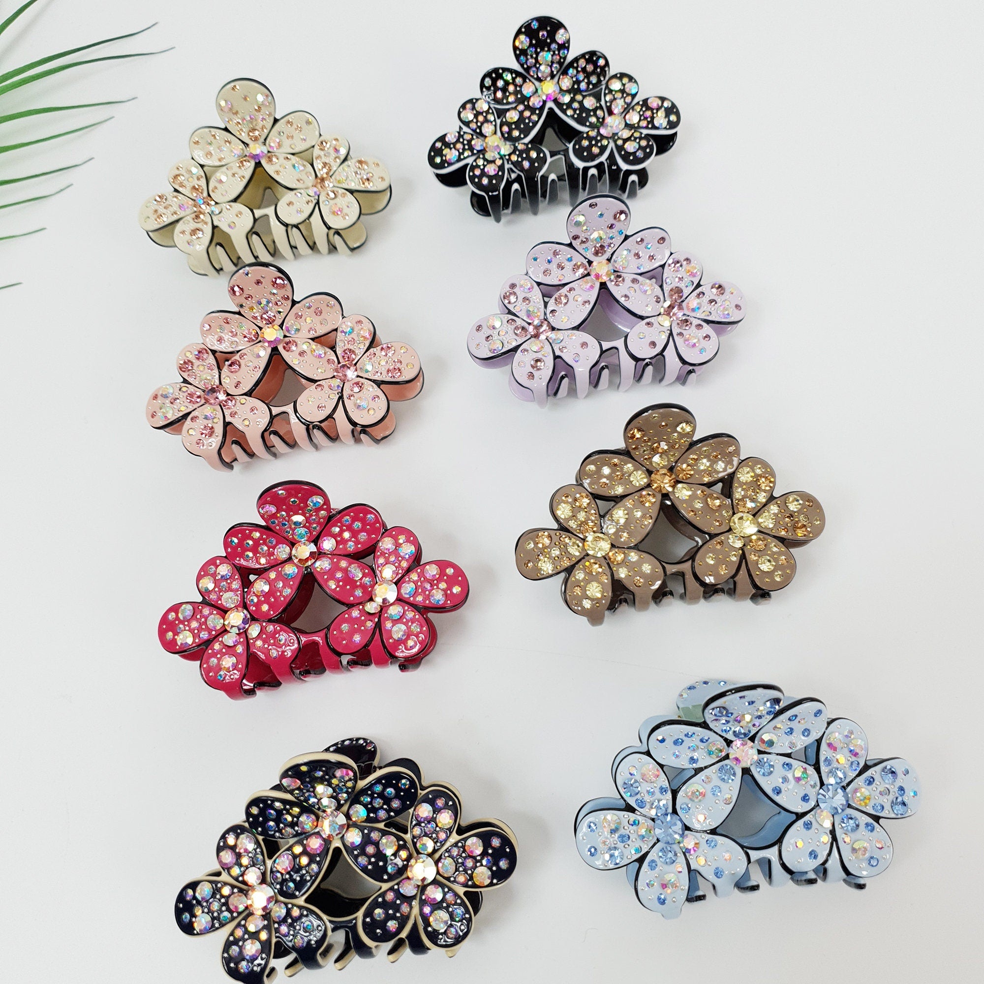 Sparkling crystal flower hair claw clip in pastel acetate, elegant triple blossom design for half-up hairstyles, 3 inch jaw clip for women. Eight hair clips with different colored bases and rhinestones. The clips are shaped like three flowers with rhinestones. The colors of the clips are black, pink, brown, red, blue, and white.