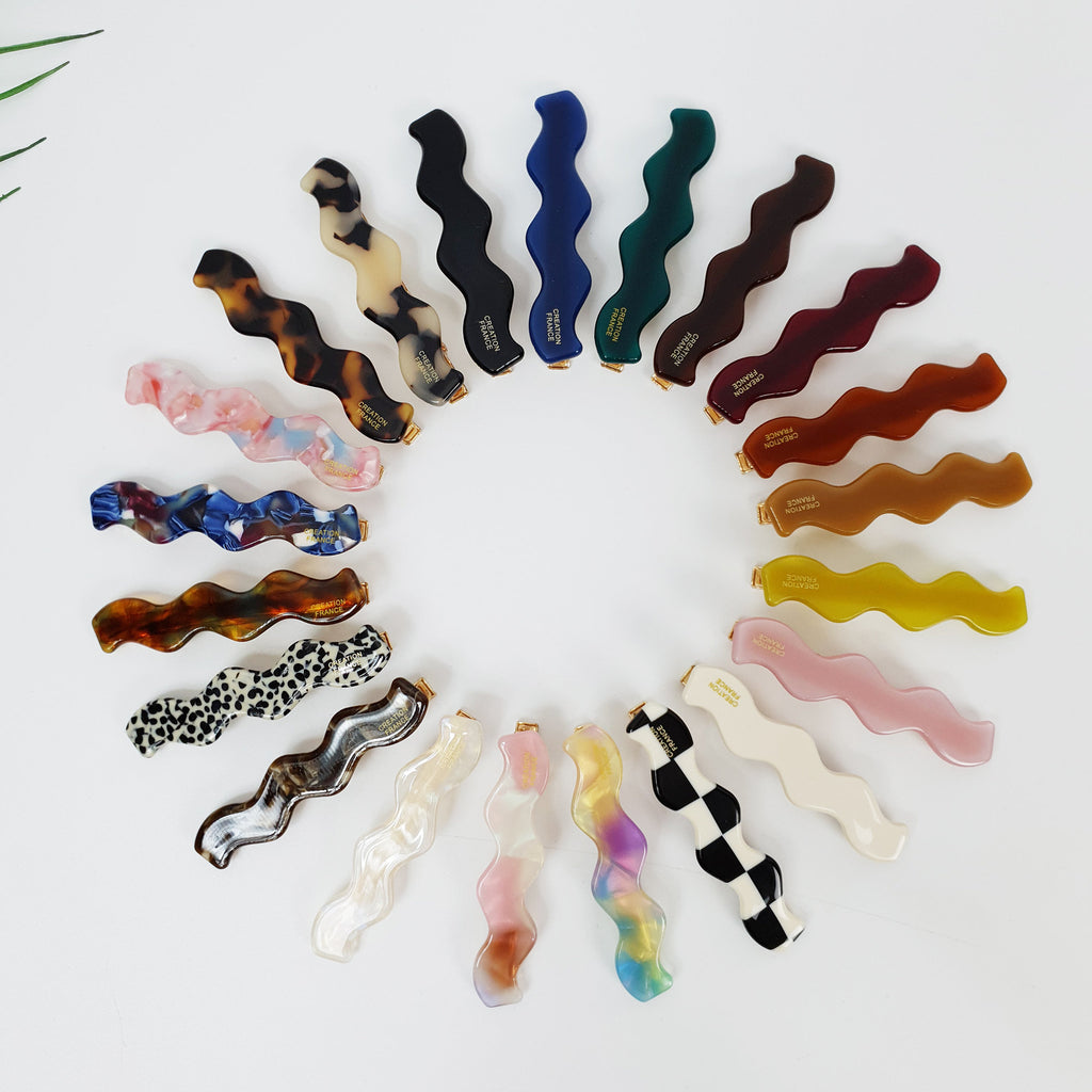 Wavy cellulose acetate hair barrette in tortoise and pastel colors, 2.6 inch squiggle shape alligator clamp for side bangs, trendy minimalist styling tool. A collection of 18 wavy hair clips in various colors and patterns including brown, black, white, pink, blue, green, yellow, and leopard print. The clips are arranged in a circle on a white background.