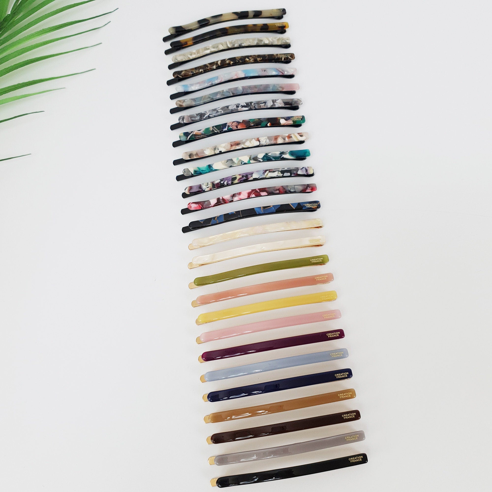 Long cellulose acetate bobby pins in tortoise shell, marble, and solid colors. Minimalist 3.5 inch hair slides for buns and side swept hairstyles. Glossy vintage style hair accessories for women. A set of twenty hair clips in various colors and patterns. The clips are made of plastic and have a gold metal clasp. The clips are arranged vertically in a row, with the top clip being a brown and black patterned clip.