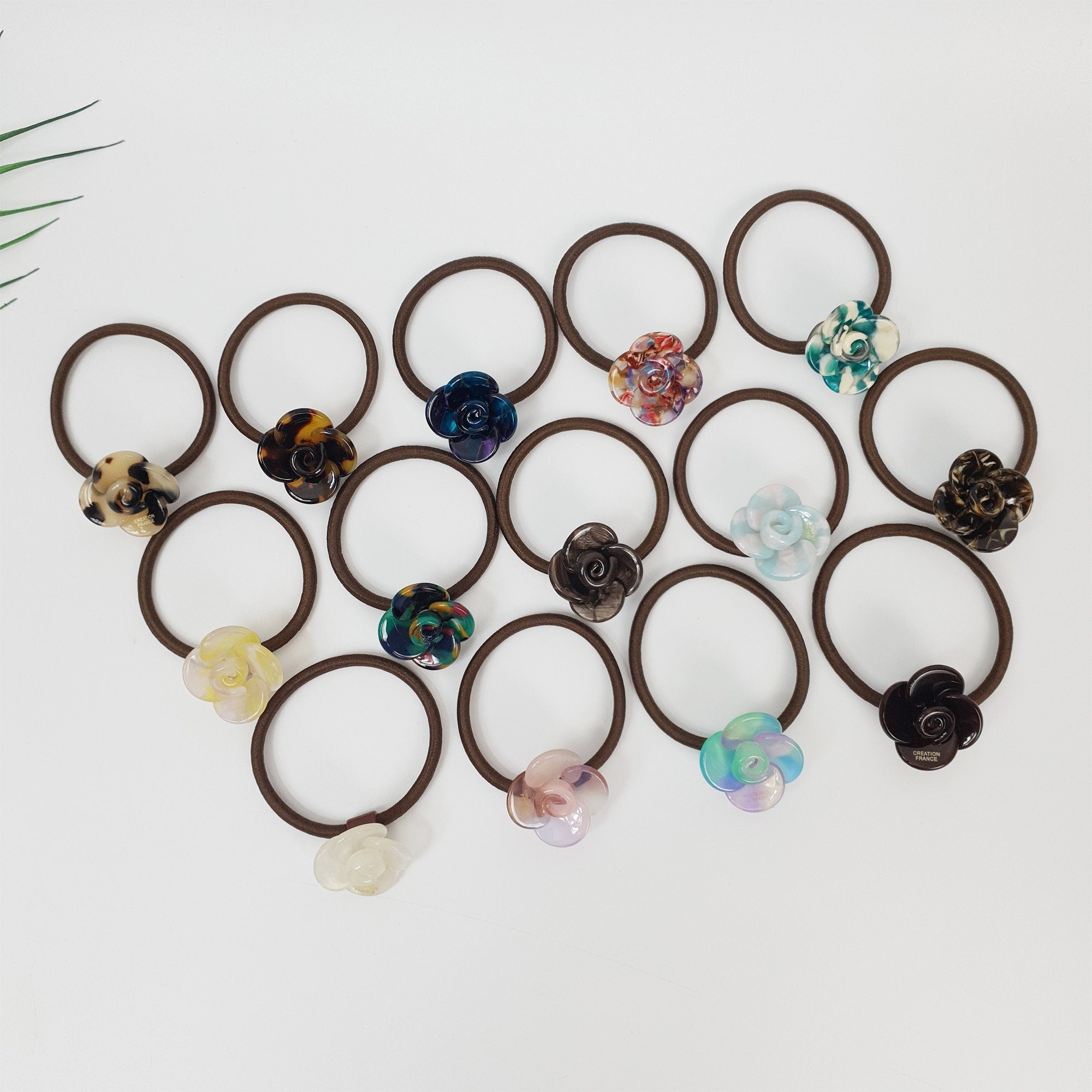 Acetate flower hair tie with camellia design. Tortoise shell and marble pattern rose ponytail holder. Cute Korean hair accessory for women.  A collection of brown hair ties with various colored flower embellishments. The flowers are made of plastic and have a variety of patterns and colors, including blue, green, pink, purple, and brown.