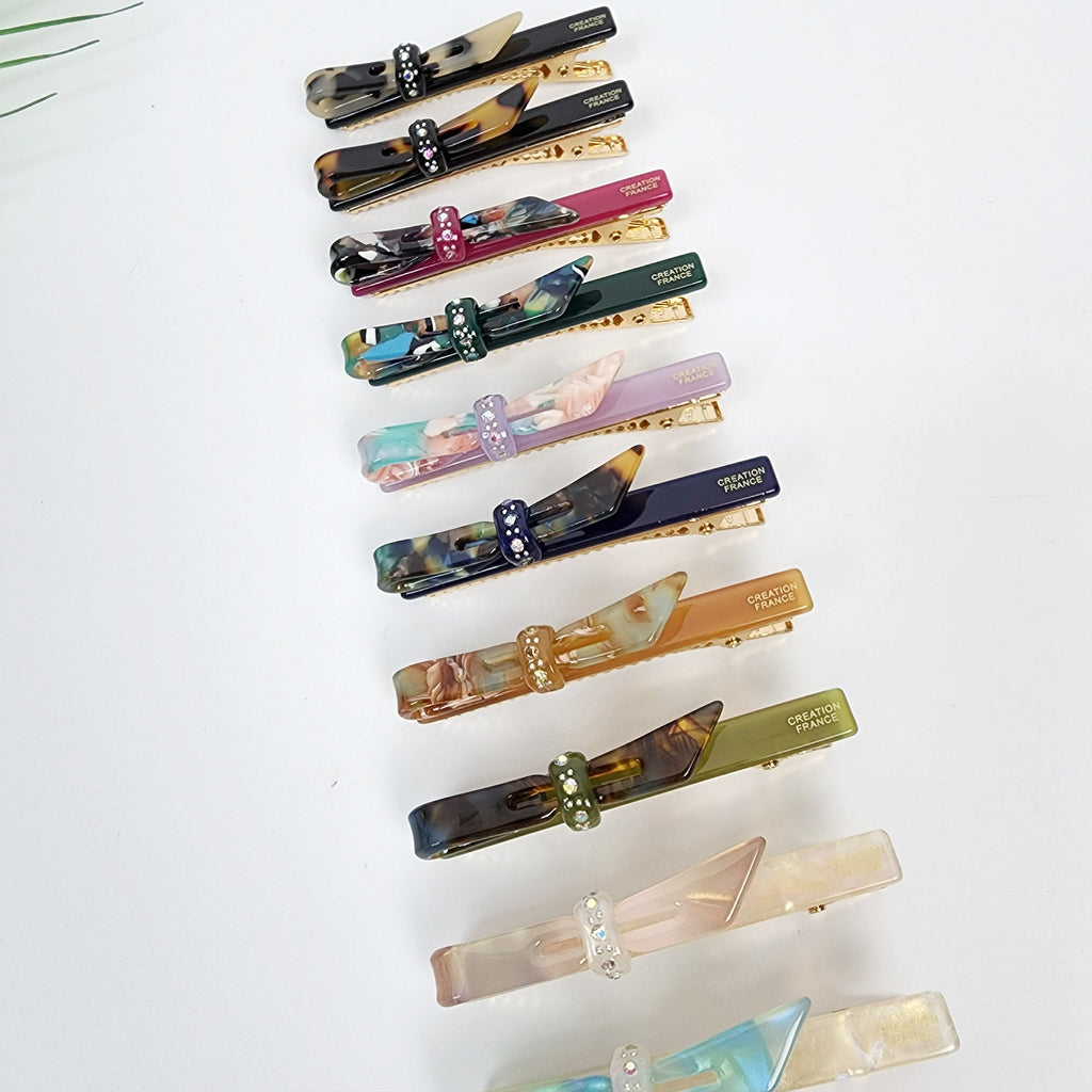 Small cellulose acetate bow hair clip with rhinestone detail. Tortoise shell and colorful alligator clips for bangs. 2.6 inch side hair pin for women and girls. A set of twelve hair clips with bows. The bows are made of different colored and patterned plastic. Each bow has a rhinestone in the center. The clips are gold colored and have the words 'CREATION FRANCE' printed on them.