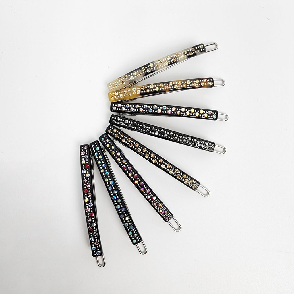 Cellulose acetate thin hair clip with rhinestones. Small 2 inch sparkling snap barrette for bangs and side hair styling. A set of eight black hair clips with rhinestones. The clips are arranged in a semi-circle, with the tips of the clips pointing towards the center. The rhinestones are different colors, including clear, gold, and multi-colored.