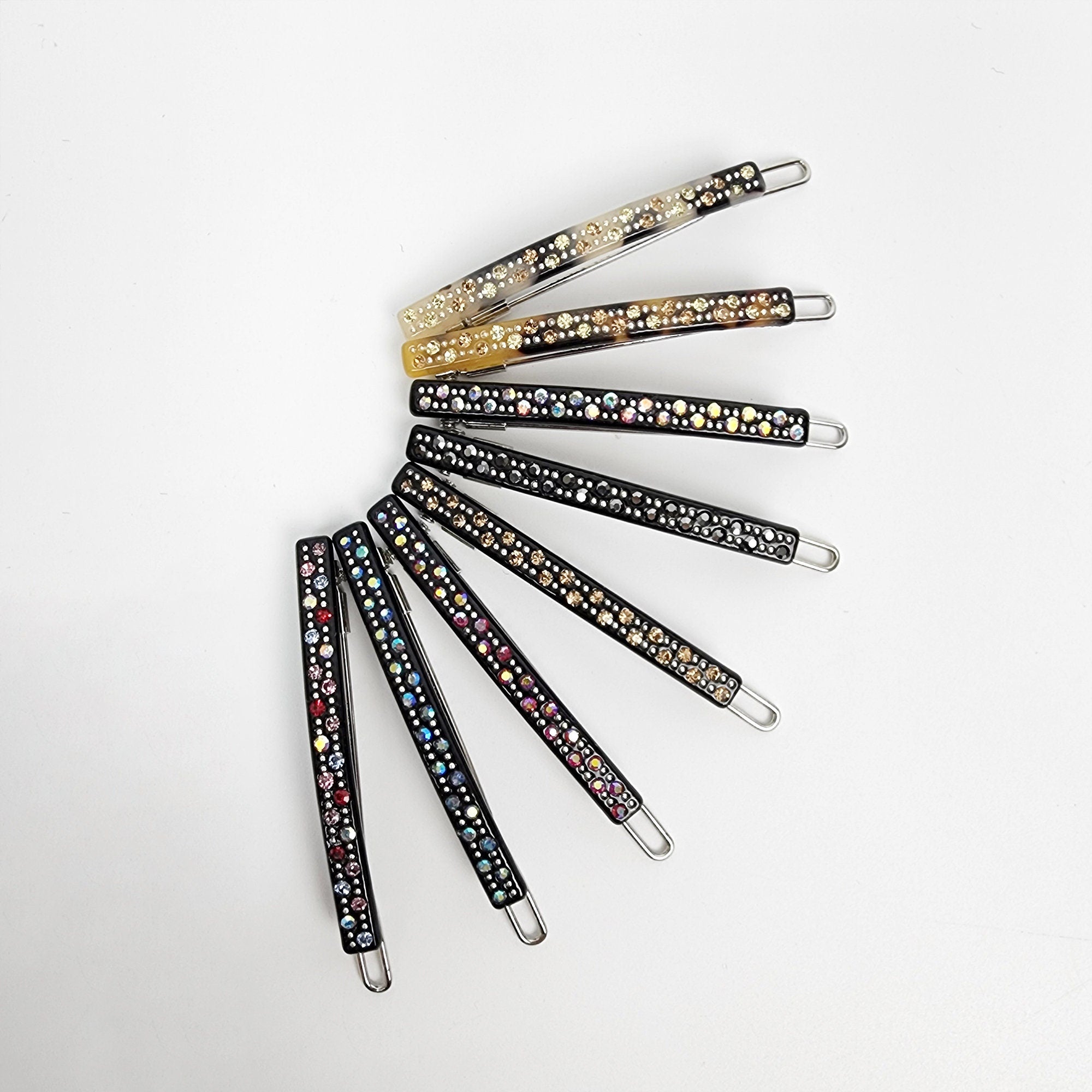 Cellulose acetate thin hair clip with rhinestones. Small 2 inch sparkling snap barrette for bangs and side hair styling. A set of eight black hair clips with rhinestones. The clips are arranged in a semi-circle, with the tips of the clips pointing towards the center. The rhinestones are different colors, including clear, gold, and multi-colored.