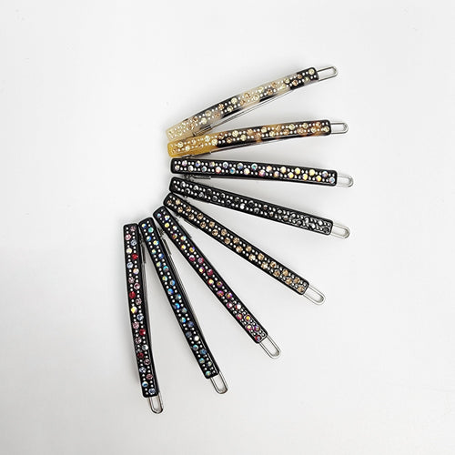 Cellulose acetate thin hair clip with rhinestones. Small 2 inch sparkling snap barrette for bangs and side hair styling. A set of eight black hair clips with rhinestones. The clips are arranged in a semi-circle, with the tips of the clips pointing towards the center. The rhinestones are different colors, including clear, gold, and multi-colored.