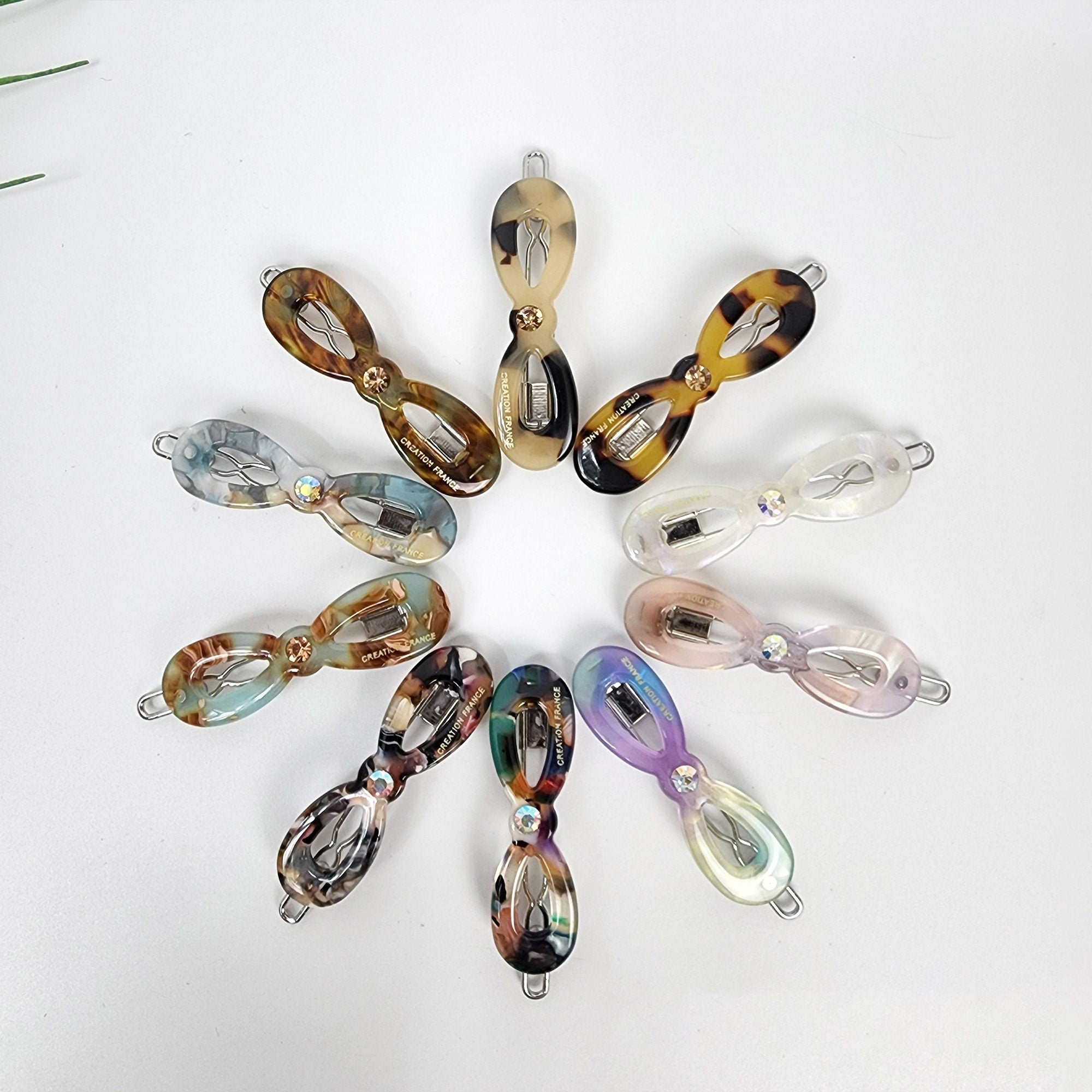 Cellulose acetate mini infinity ribbon hair clip with crystal accent, small tortoise shell snap barrette for bangs, cute Korean hair accessories. Ten hair clips in various colors and patterns, including brown, white, blue, green, and purple. The clips are all in the shape of a bow and have a small rhinestone in the center. The clips are arranged in a circle on a white background.