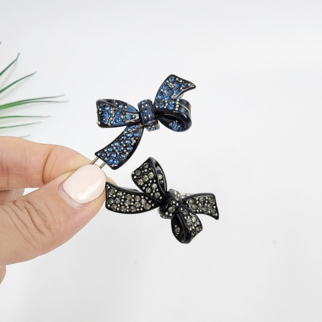 Cellulose acetate ribbon clip with rhinestones. Small sparkly snap barrette in black and navy for elegant side hair styling. Two black jeweled bow hair clips. One bow is blue and the other is gray. The bows are both made of rhinestones and have a metal clip on the back.
