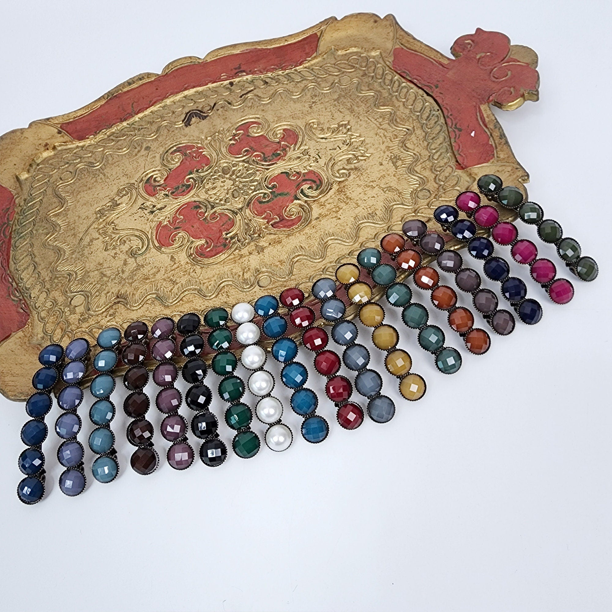 Vintage style antique bronze hair barrette with six round faceted resin stones, elegant french clip accessory for half-up hairstyles or side bangs. A set of 14 hair clips with a variety of colored glass beads. The beads are arranged in a cascading pattern on a gold tray with red and gold accents. The beads are in shades of blue, green, red, brown, and yellow.