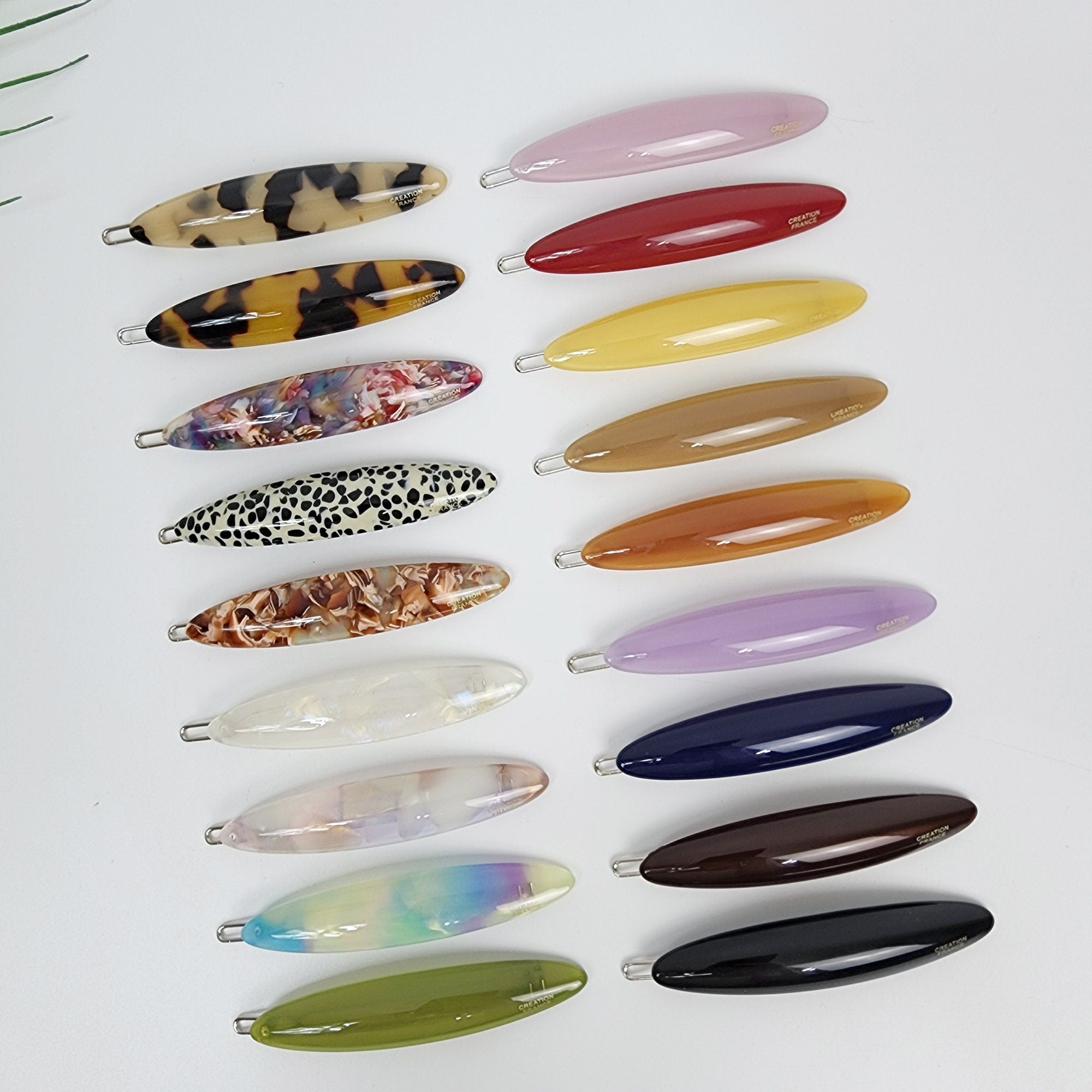 Cellulose acetate oval hair barrette featuring tortoise shell patterns and pastel colors for women and girls styling side bangs. A collection of 15 colorful hair clips. The clips are made of plastic and have a variety of patterns and colors, including brown, black, white, yellow, orange, red, purple, green, and blue. The clips are arranged in two rows, with the top row having 8 clips and the bottom row having 7 clips.