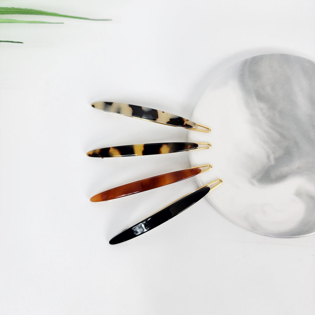 Cellulose acetate super slim long oval hair clips in tortoise shell and black, minimalist snap barrette for bangs and side hair styling. Four hair clips in different colors and patterns. The clips are made of plastic and have a gold metal clasp. The clips are arranged in a row on a white surface. The clips are black, brown, white and black, and brown and white.