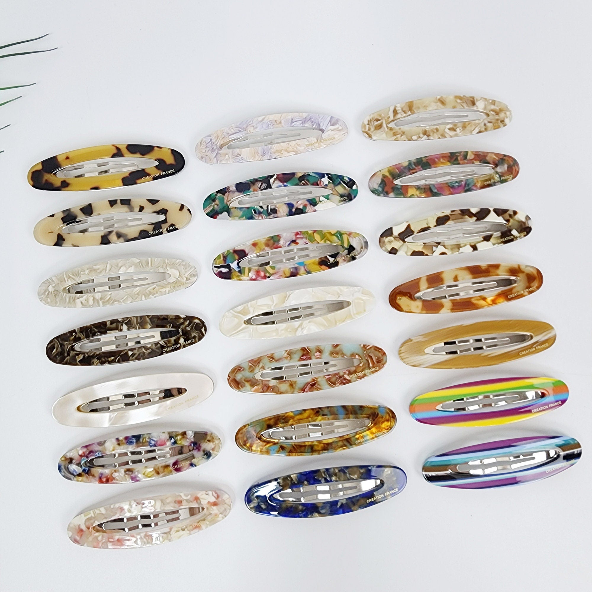 Elongated cellulose acetate snap clip in tortoise and art patterns, 3.3 inch large oval barrette for thick bangs, retro minimalist hair accessory. Twenty assorted oval-shaped hair clips with different patterns and colors. The clips are made of plastic and have a silver metal bar in the center. The patterns include leopard print, floral, striped, and abstract designs. The colors include brown, white, black, blue, yellow, green, and purple.