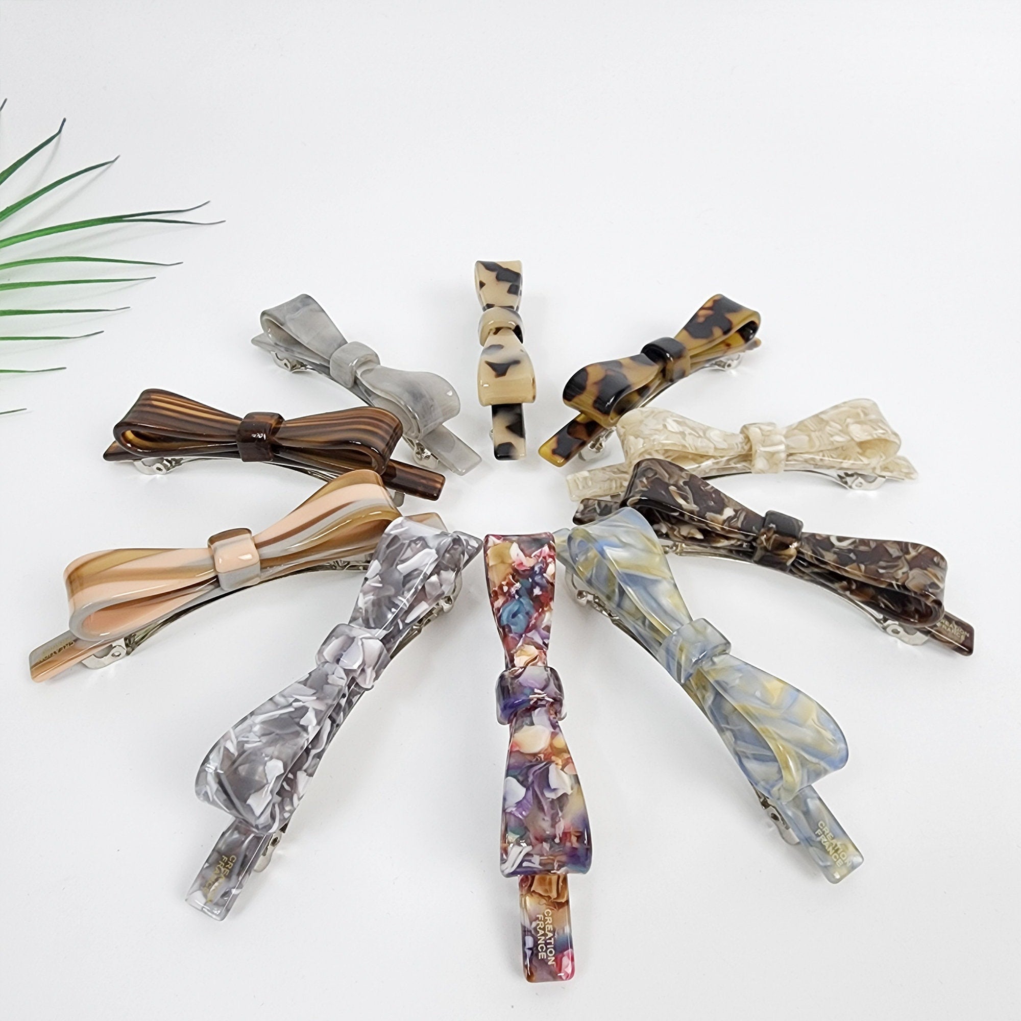 Slim bow tie shaped cellulose acetate hair barrette in tortoise shell and marble patterns. Automatic spring clip for ponytail or half-up styling, preppy office hair accessory. A collection of ten assorted hair clips with bow designs. The bows are made of various materials including brown, beige, white, and multi-colored patterned plastic. The clips are all different sizes and shapes.