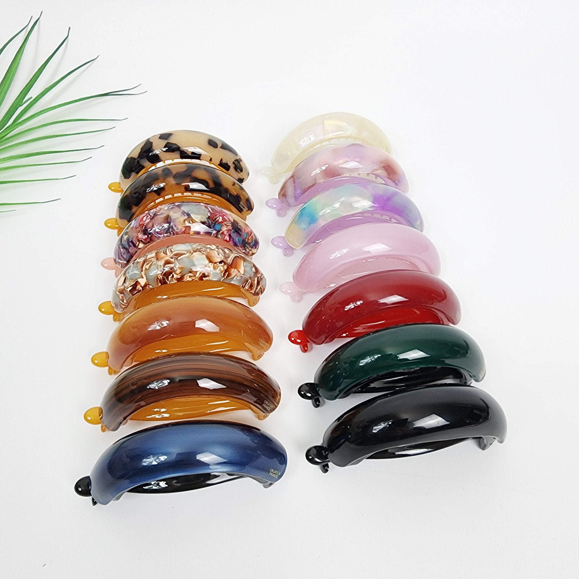 Cellulose acetate banana clip in tortoise shell and solid colors. Curved ponytail holder 3 inch size for secure half updos. A set of 14 hair clips in various colors and patterns. The clips are all made of plastic and have a curved shape. The colors include brown, black, blue, green, red, pink, and white. The patterns include leopard print, floral, and abstract designs.