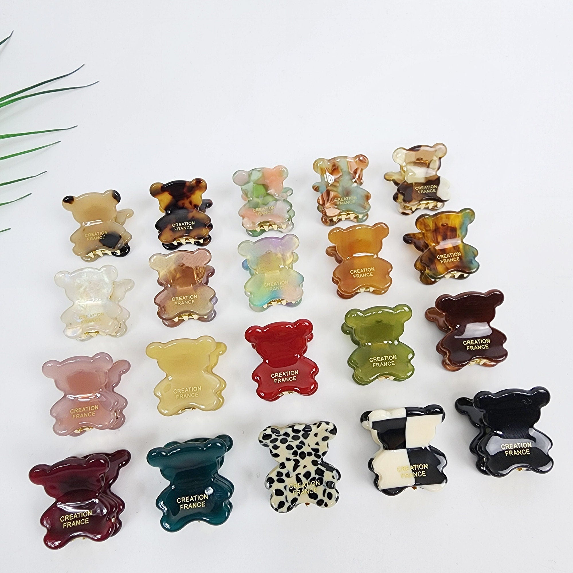 Cellulose acetate bear hair claw clip in tortoise shell and checkerboard patterns, cute mini animal hair accessories for toddler girls and bangs styling. A collection of 19 bear-shaped hair clips in various colors and patterns, including clear, pink, red, green, brown, black, white, and multi-colored. The clips are all made of plastic and have a gold-colored inscription that reads 'CREATION FRANCE'.