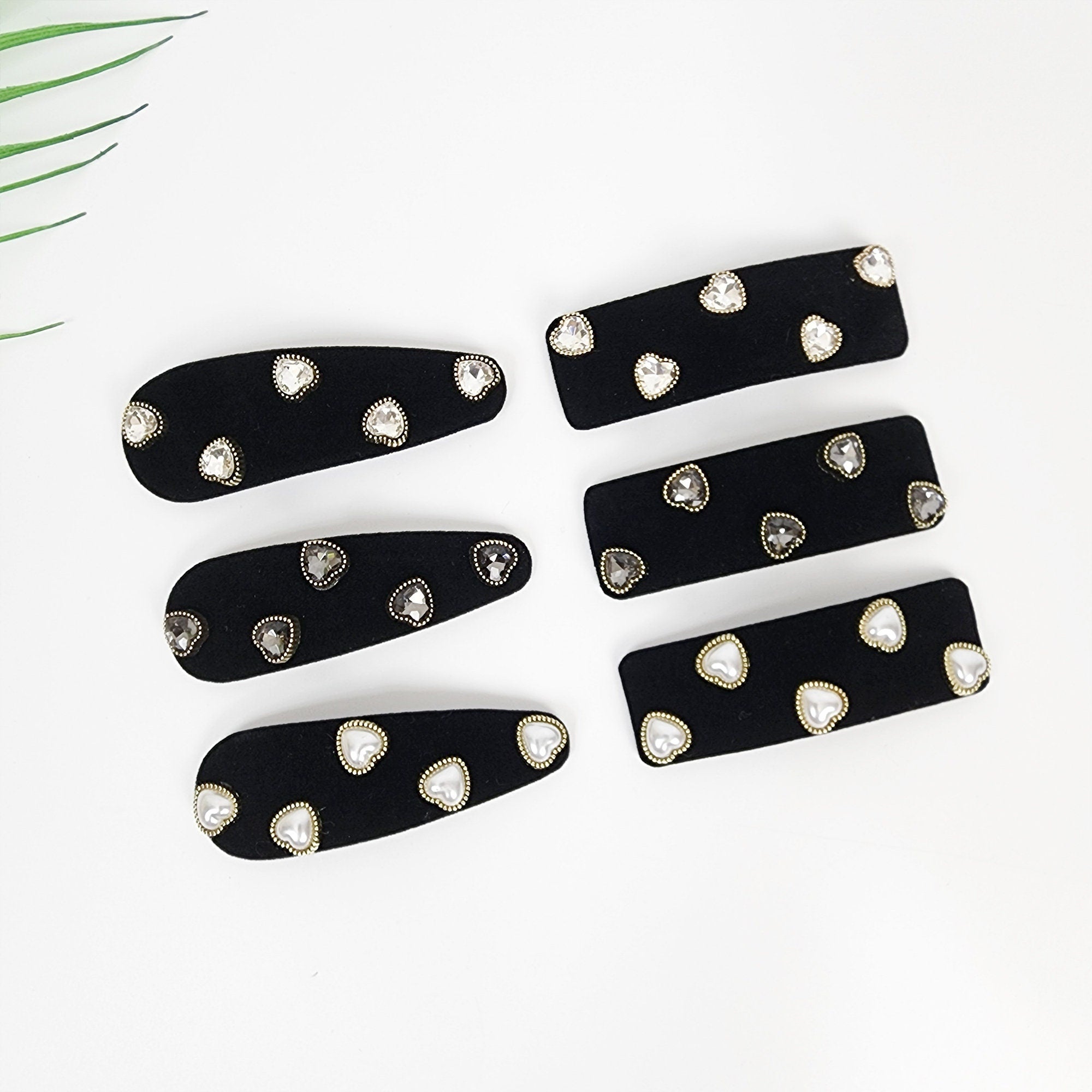 Black velvet snap hair clip with heart shaped pearls and rhinestone details for side bangs styling.  Five black velvet hair clips with gold trim and clear rhinestone hearts. The clips are arranged in two rows, with three clips in the top row and two clips in the bottom row.