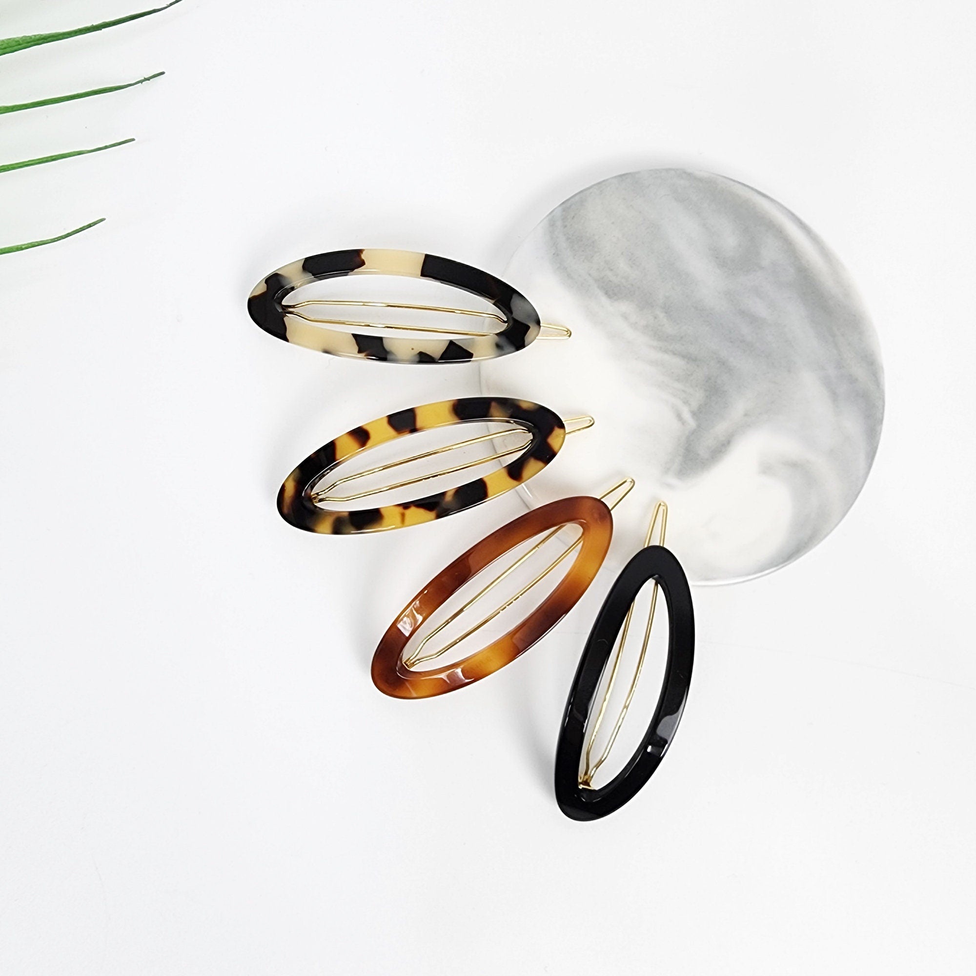 Cellulose acetate oval ring snap hair clip in tortoise shell pattern. 2.4 inch minimalist french style barrette for women holding side bangs or flying hair.  Five oval-shaped hair clips with gold metal accents. The clips are made of brown and black tortoise shell patterned plastic and solid black plastic. The clips are arranged on a white marble surface.