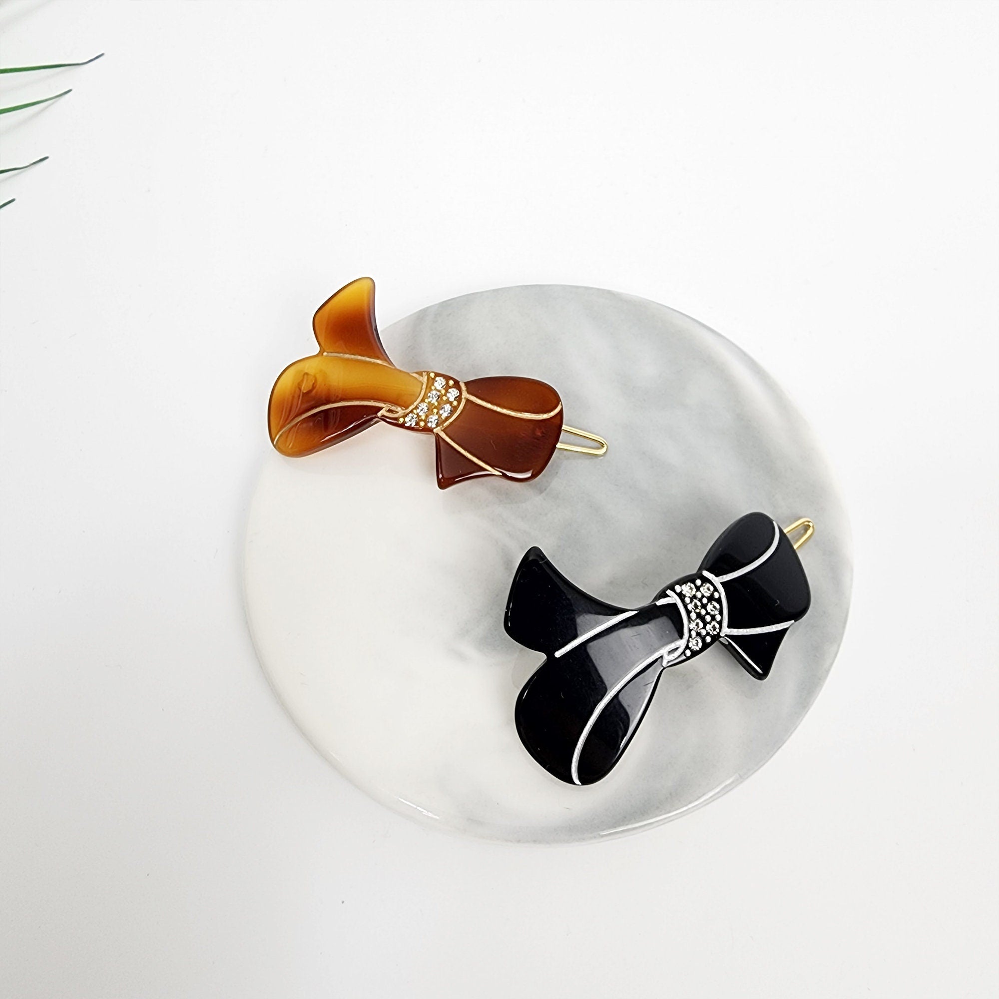 Cellulose acetate bow hair clip with crystal accents. Small 1.8 inch snap barrette for bangs and side hair available in tortoise and black. Two hair clips in the shape of bows. One bow is brown with rhinestones and the other is black with rhinestones.