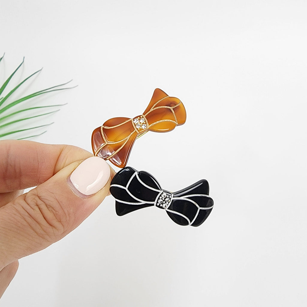 Cellulose Acetate Bow Snap Clip with Crystal Accents Small Ribbon Barrette for Bangs Tortoise Shell Pattern Korean Hair Accessory Two hair clips shaped like bows. One bow is brown with a clear rhinestone in the center. The other bow is black with a clear rhinestone in the center.
