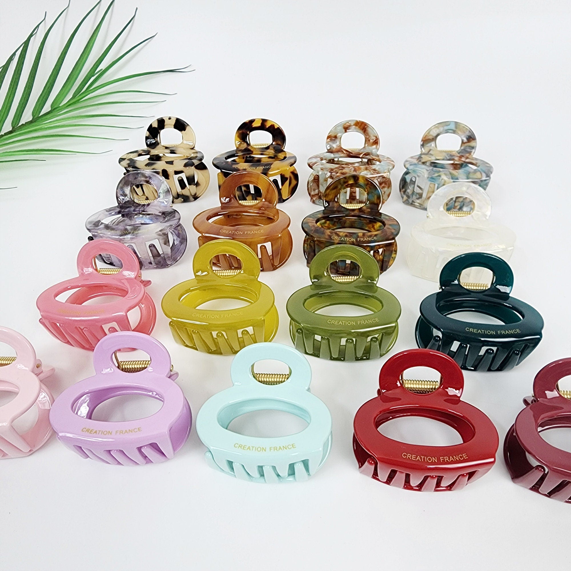 Small 2.2 inch cellulose acetate hair claw with snowman shape and U-wire spring. Rounded jaw clip for half-up hairstyles available in tortoise and pastel colors. A collection of 15 colorful plastic hair clips. The clips are all oval shaped with a double loop design. The clips are in various colors including pink, purple, green, red, blue, brown, and white. The clips have the words 'Creation France' printed on them.