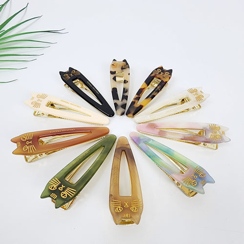 Cat shaped cellulose acetate hair slide with gold whiskers details. Tortoise and marble pattern alligator clip for side bangs, kawaii hair accessory. Ten cat-shaped hair clips in various colors, including black, brown, beige, green, pink, and purple. Each clip has a gold metal clasp and a gold cat face design with the words 'CREATION FRANCE' printed on the back.