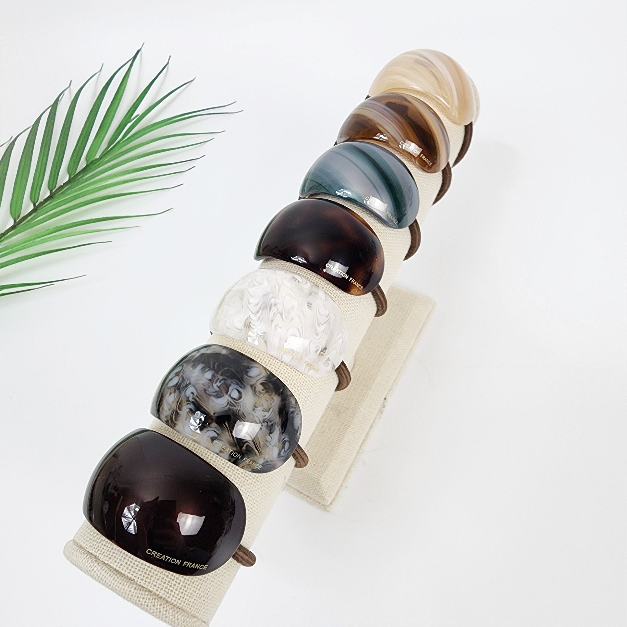 Glossy acetate curved hair tie in beige and brown marble pattern. Minimalist pony cuff for thick hair. Korean elegant hair accessory for women. Seven hair scrunchies displayed on a white stand. The scrunchies are made of different materials and colors, including brown, white, gray, and beige. The scrunchies are all oval shaped and have a glossy finish. The text 'CREATION FRANCE' is printed on the bottom scrunchie.