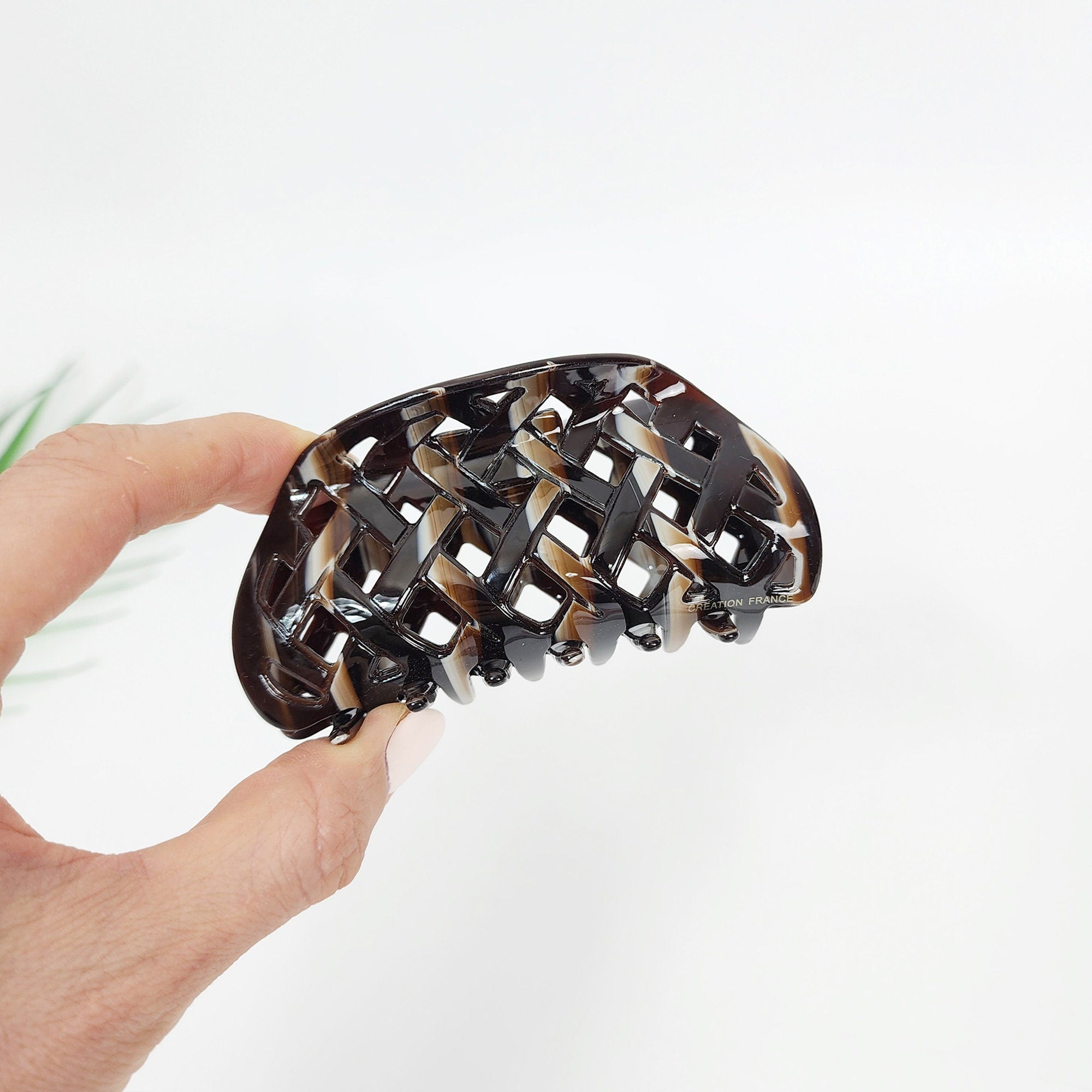 Medium cellulose acetate hair claw with a checkered lattice cutout design. 3.7 inch curved jaw clip in tortoise and pearlescent colors for french twist or half-up styling. A brown and black plastic hair claw with a lattice design. The claw is designed to hold hair in place.