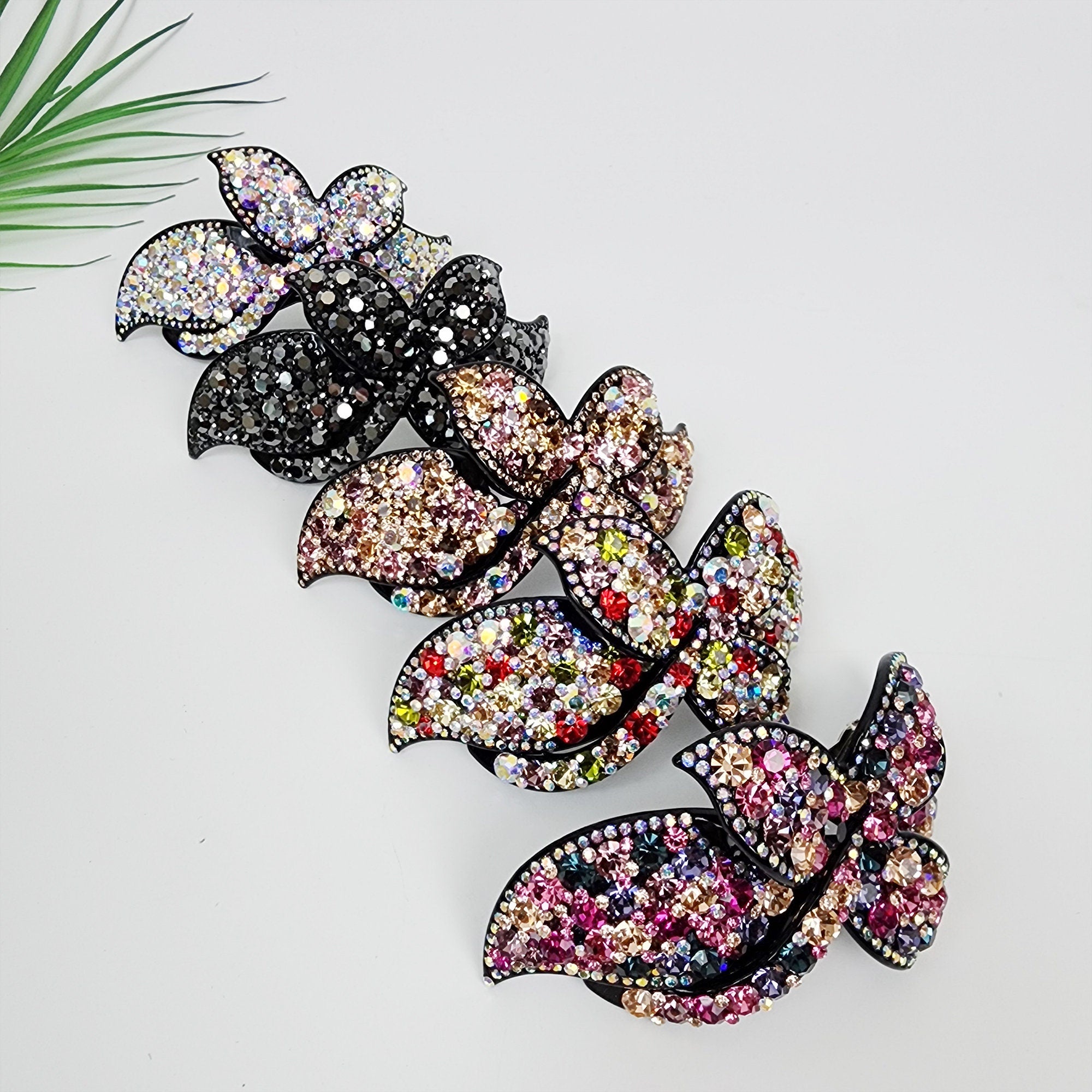 Sparkling rhinestone French barrette with large crystal bow design, elegant acetate hair clip for formal updos, weddings, and party styles. Three black hair clips with rhinestones in different colors. The top clip has clear rhinestones, the middle clip has pink and gold rhinestones, and the bottom clip has pink, purple, and blue rhinestones.