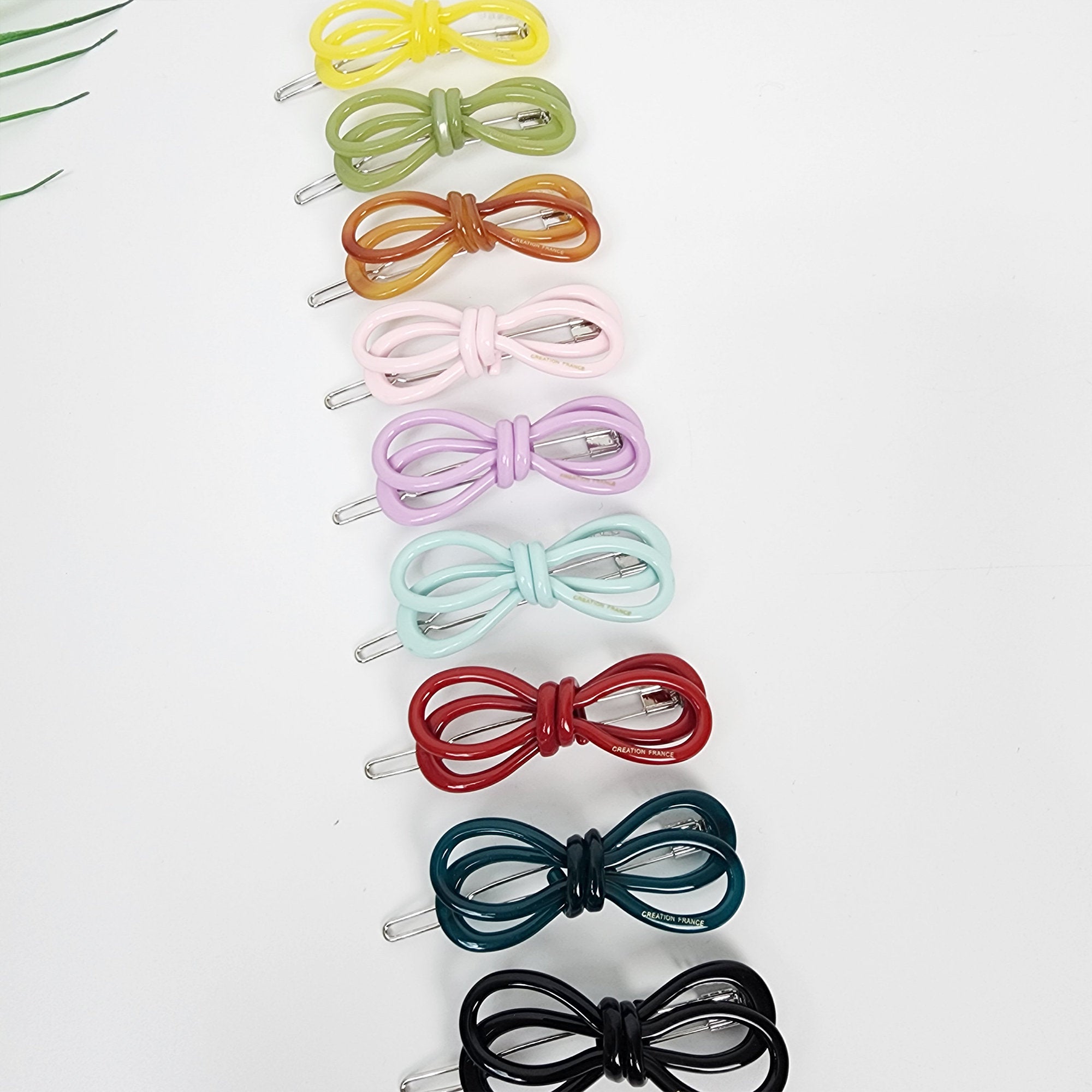 Cellulose acetate twin loop bow hair clip in pastel and solid colors mini snap barrette for side bangs.  A set of ten hair clips with bow designs in various colors including yellow, green, brown, pink, purple, blue, red, teal, and black. The clips are arranged vertically in a row from top to bottom.