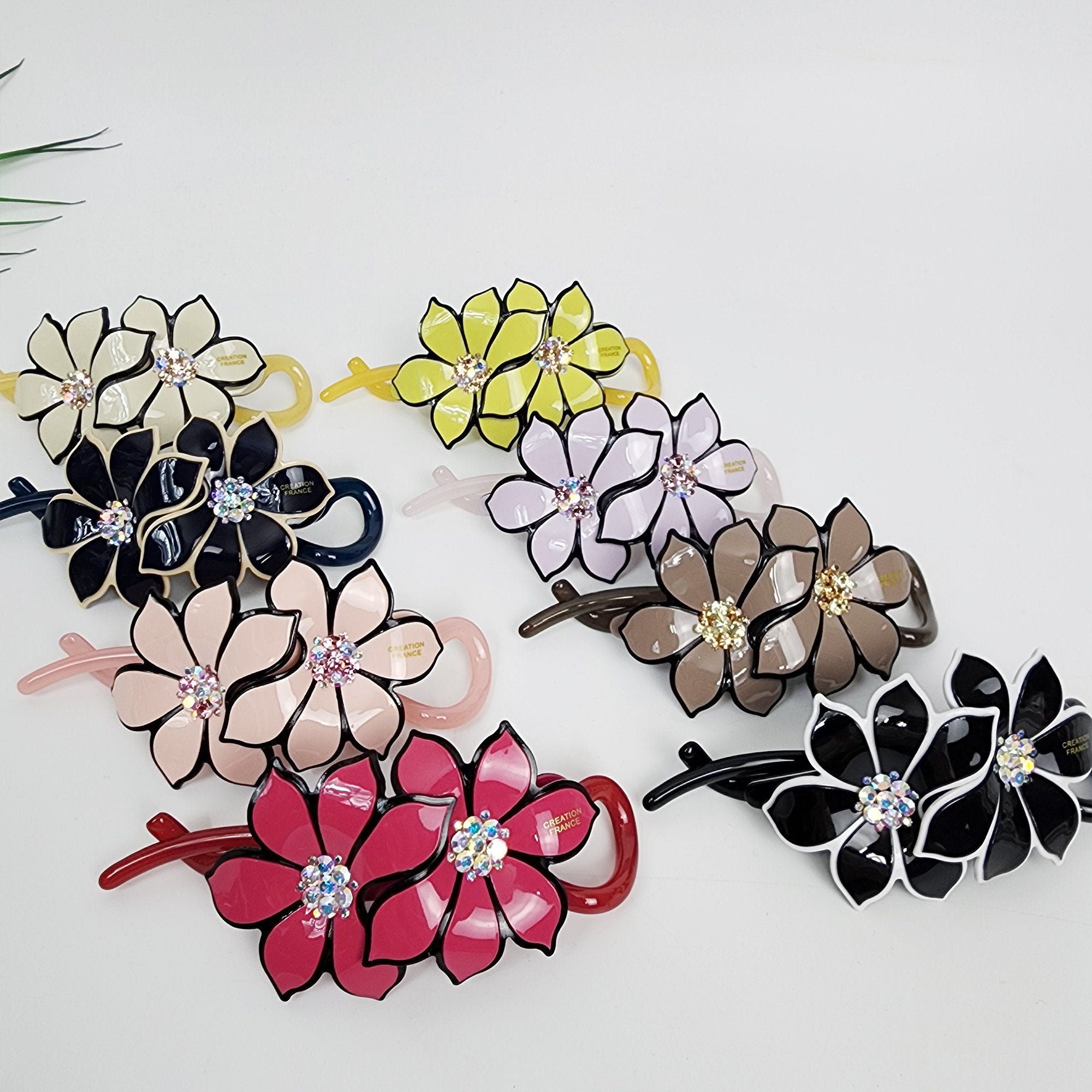 Cellulose acetate floral banana clip with rhinestone center double flower hair accessory for women half updo styling Assortment of colorful flower-shaped hair clips with rhinestones. The clips are made of plastic and have a variety of colors, including pink, yellow, black, and brown. The clips are designed to hold hair in place and add a touch of style to any hairstyle.