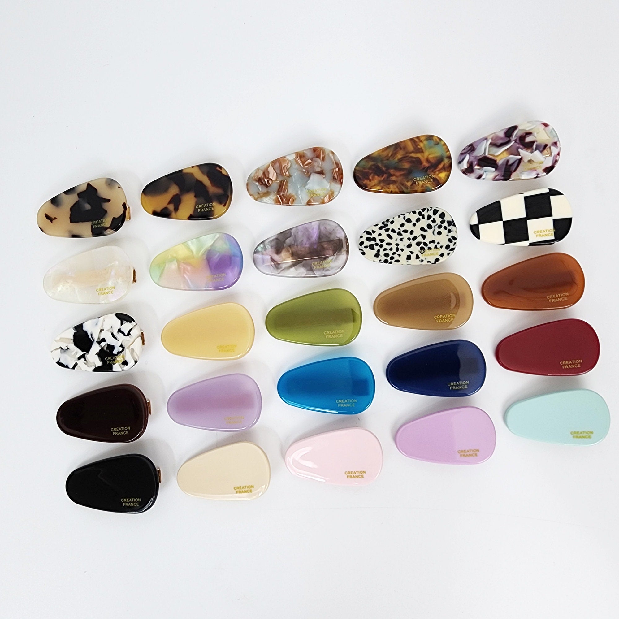 Cellulose acetate flat triangle hair clip tortoise and checkerboard patterns small alligator slide for bangs. A collection of twenty-two assorted plastic hair clips in various colors and patterns. The clips are all shaped like a teardrop and have a gold metal clasp. The patterns include tortoise shell, leopard print, checkered, marble, and solid colors. The clips are all labeled 'Creation France'.