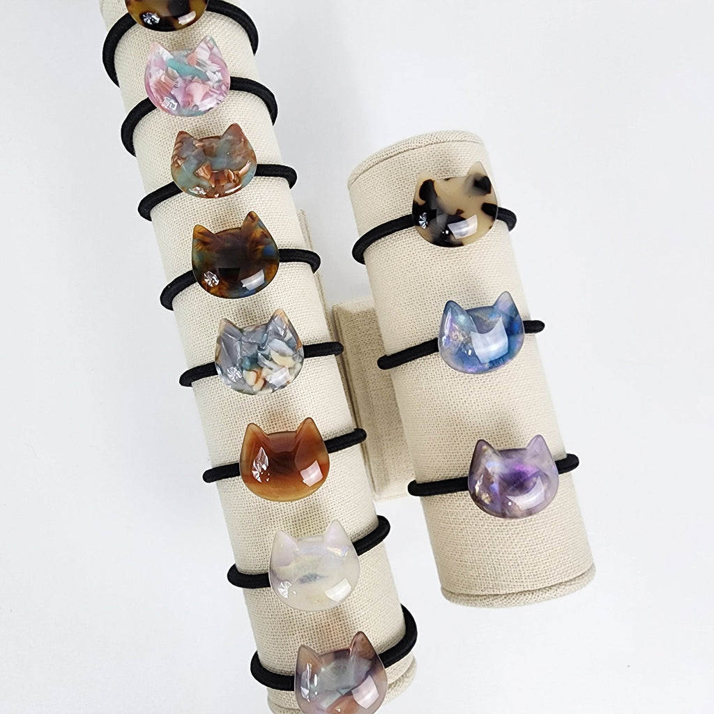Cellulose acetate cat hair tie in tortoise shell and marble patterns. Cute kitten shaped ponytail holder for women and girls with a glossy finish. Two sets of hair ties with cat face charms. The hair ties are black and the charms are made of translucent plastic with different colors and patterns. The charms are in the shape of a cat's face with a white background and a black outline.