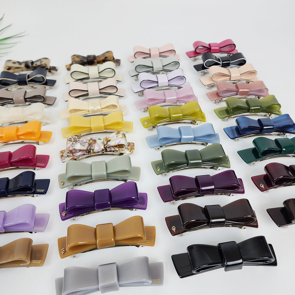 Cellulose acetate bow tie hair barrette with automatic clasp. 3.5 inch solid color ribbon hair clip for half-up hairstyles. A collection of twenty-four assorted hair clips in various colors, including purple, brown, green, blue, gray, yellow, orange, pink, and white. The clips are all in the shape of a bow with a silver metal bar in the center.