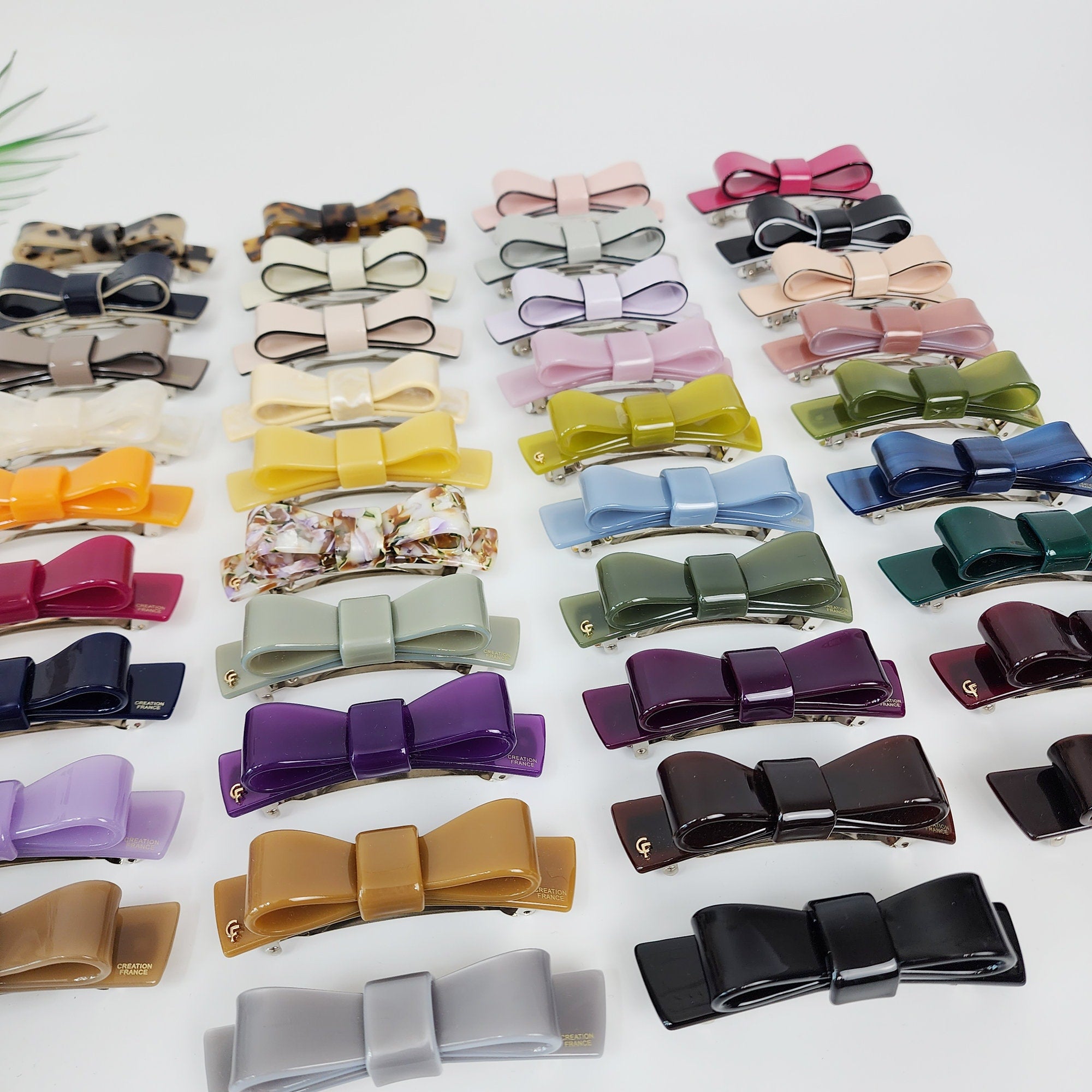 Cellulose acetate bow tie hair barrette with automatic clasp. 3.5 inch solid color ribbon hair clip for half-up hairstyles. A collection of twenty-four assorted hair clips in various colors, including purple, brown, green, blue, gray, yellow, orange, pink, and white. The clips are all in the shape of a bow with a silver metal bar in the center.