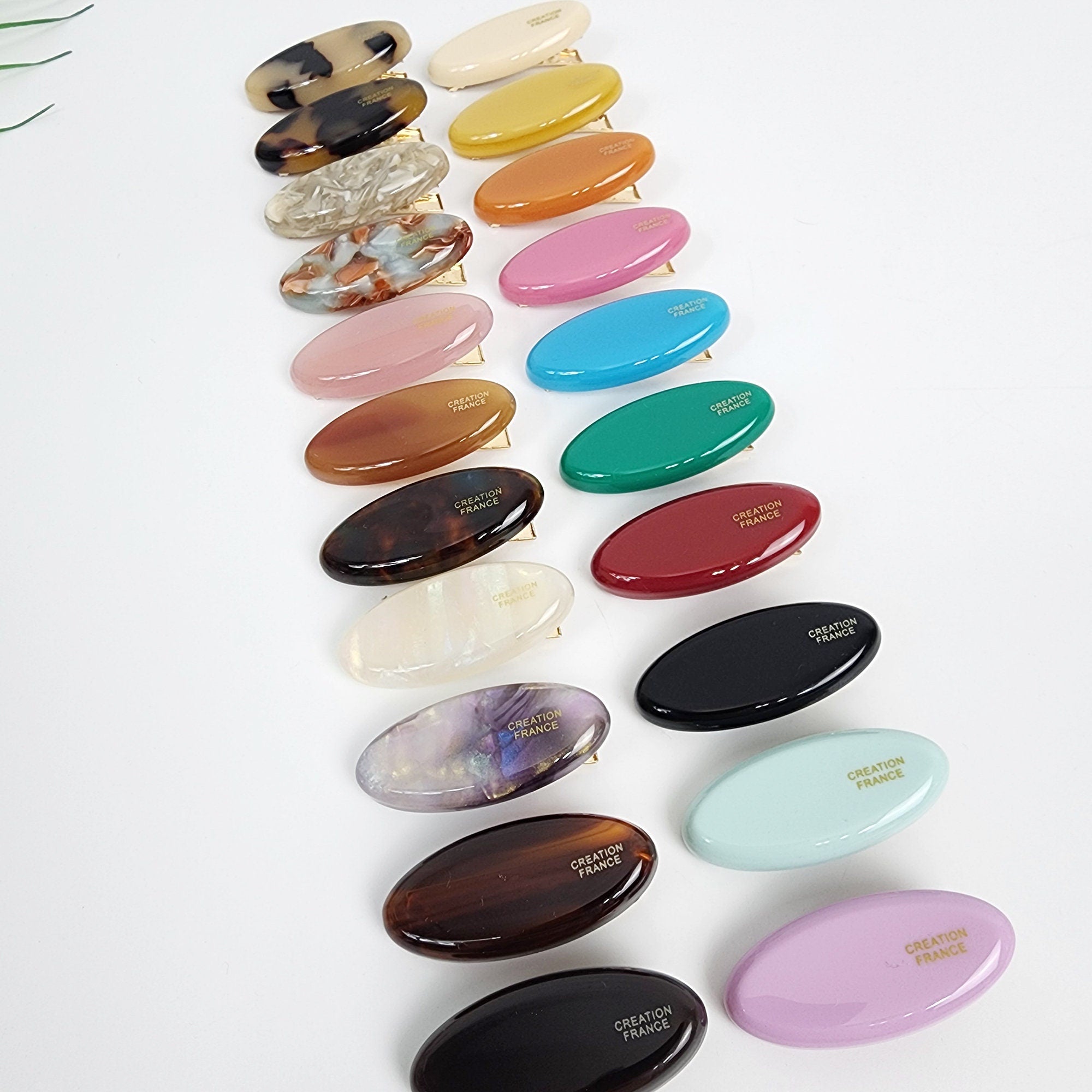 Cellulose acetate mini pebble shape hair claw clip in beige, tortoise, and pastel colors. 1.6 inch tiny oval jaw clamp for women's side bangs. Cute Korean hair accessory. A collection of 16 oval-shaped hair clips in various colors and patterns. The clips are made of plastic and have a smooth, glossy finish. Each clip has the words 'CREATION FRANCE' printed on the bottom in gold lettering.