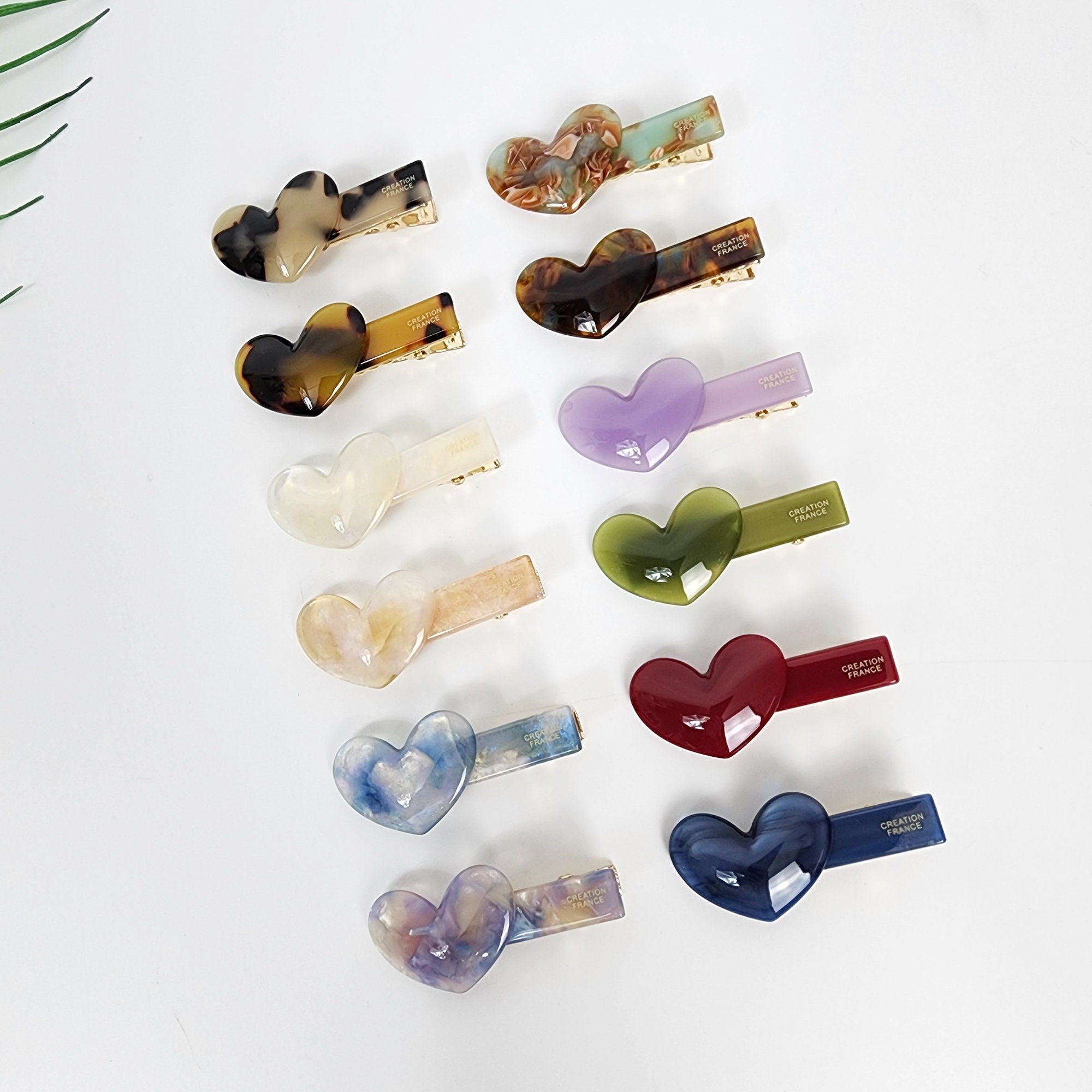 Small heart shaped cellulose acetate hair clip with alligator grip for side bangs and decorative styling. Twelve heart-shaped hair clips with gold metal clasps. The clips are made of translucent plastic and come in a variety of colors, including purple, green, red, blue, brown, and white. The clips have the words 'CREATION FRANCE' printed on them.