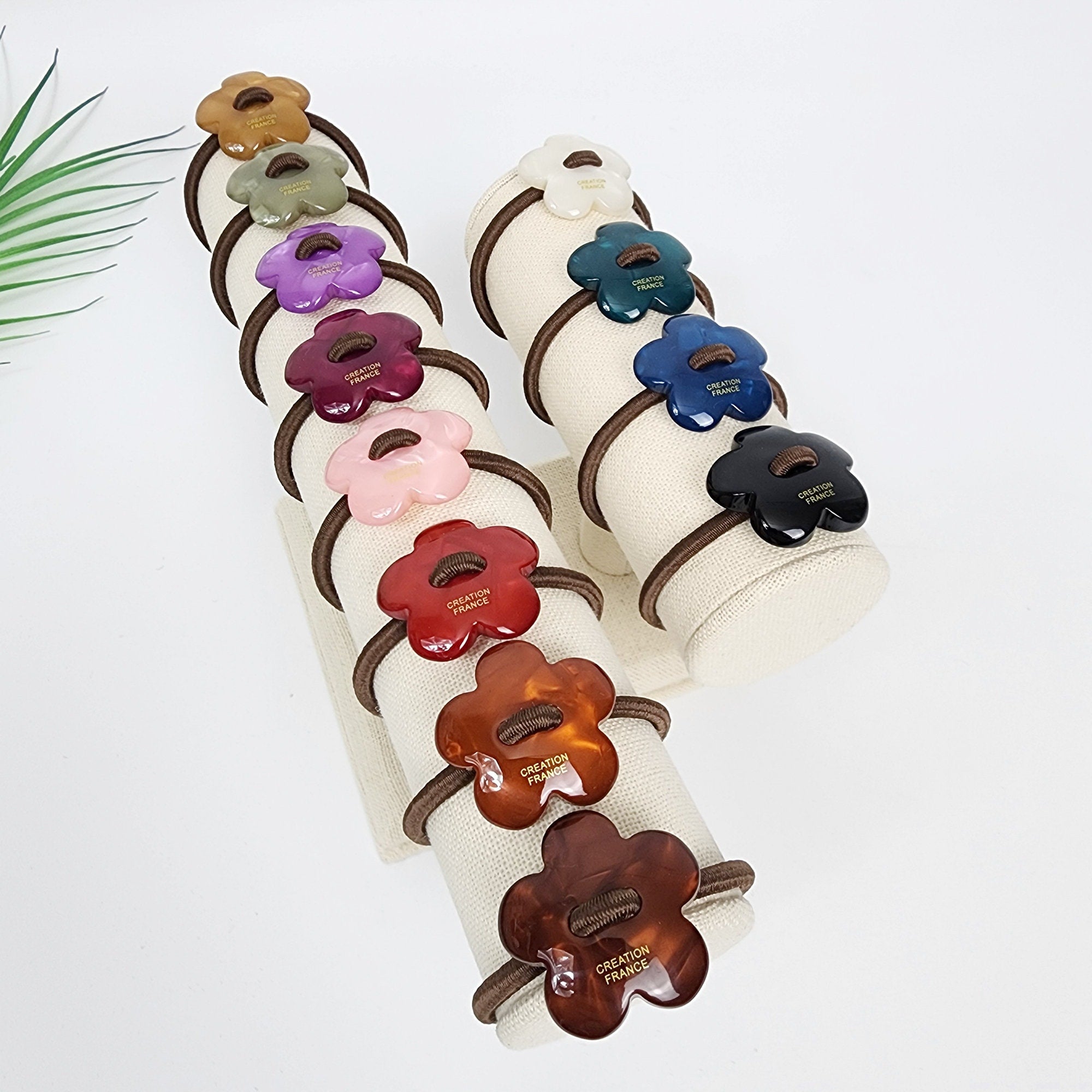 Cellulose acetate flower hair tie with stitch detail. Convex floral ponytail holder in 12 colors. Korean retro hair accessory for women. A display of 12 hair scrunchies with flower-shaped buttons. The scrunchies are arranged in two columns on a white fabric stand. The buttons are made of different colors including red, pink, brown, green, blue, black, and white. Each button has the words 'CREATION FRANCE' printed on it.