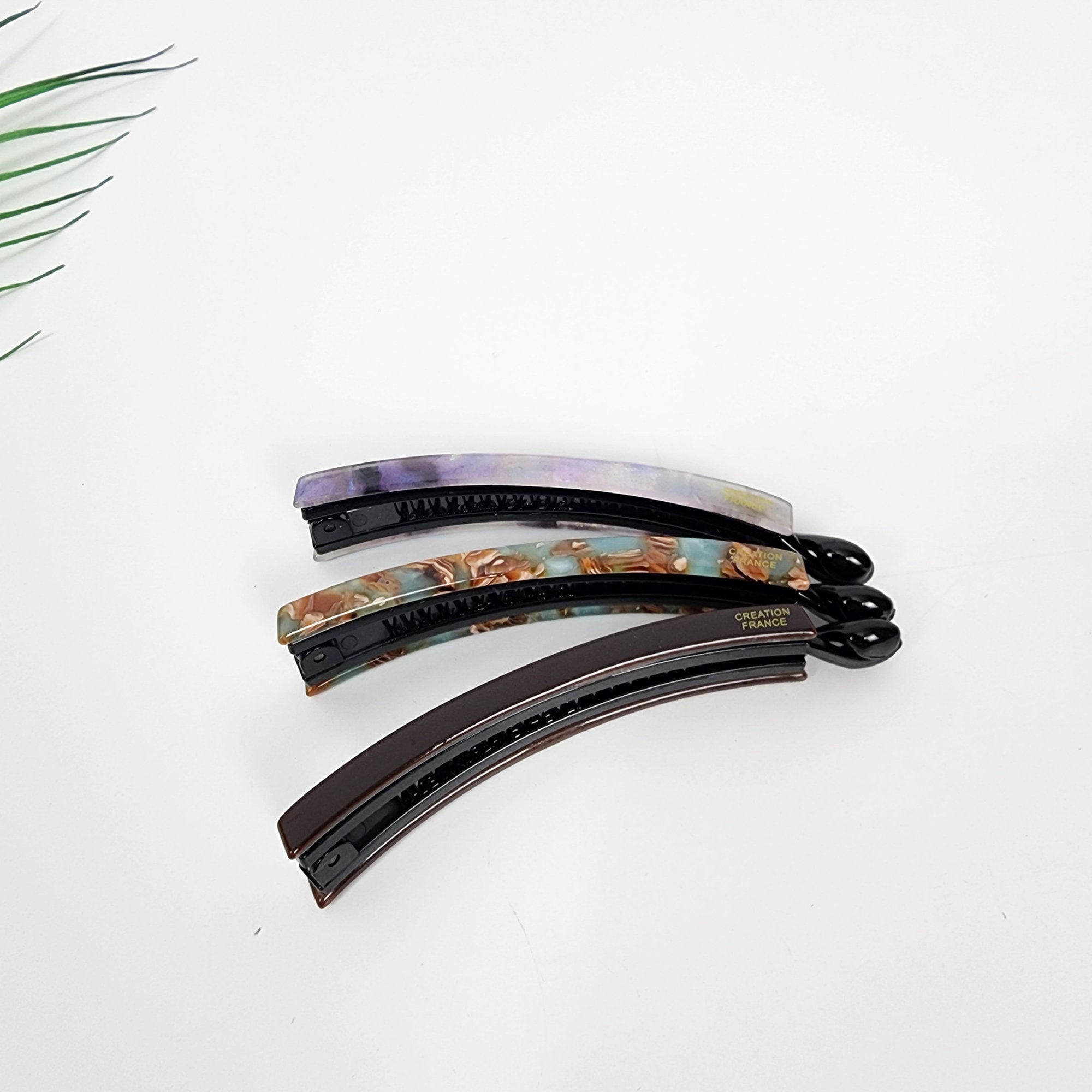 Cellulose acetate slim banana clip with inner teeth mechanism for fine hair and half updo styling. Three hair clips with a curved shape and a black base. The clips are decorated with a marbled pattern in shades of purple, brown, and blue. The bottom clip has the text 'CREATION FRANCE' printed on it.