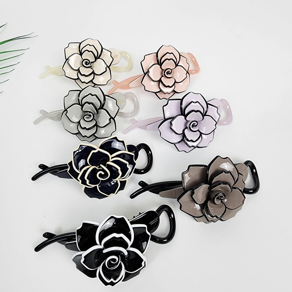 Cellulose acetate banana clip featuring a large camellia flower design with bold outlines. Elegant floral hair accessory for ponytails or half-up styles, available in black and pastel tones. Six flower shaped hair clips. The clips are made of plastic and have a black outline. The flowers are in various colors including pink, light purple, gray, brown, and black.